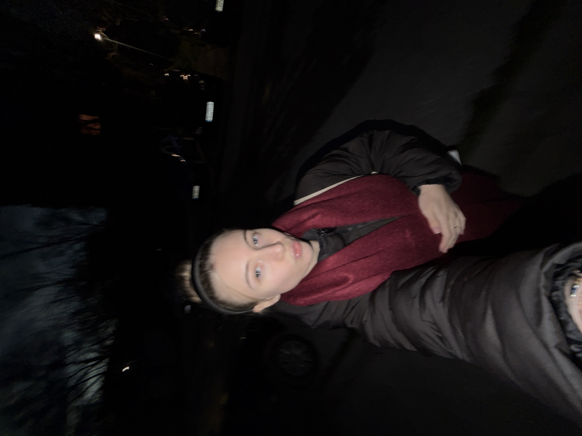 A woman stands in the city of Plovdiv, Bulgaria at night. The woman is wearing a black puffy coat and a maroon scarf. The scene is dark, with city lights visible in the background. A car wheel is visible to the left of the woman.