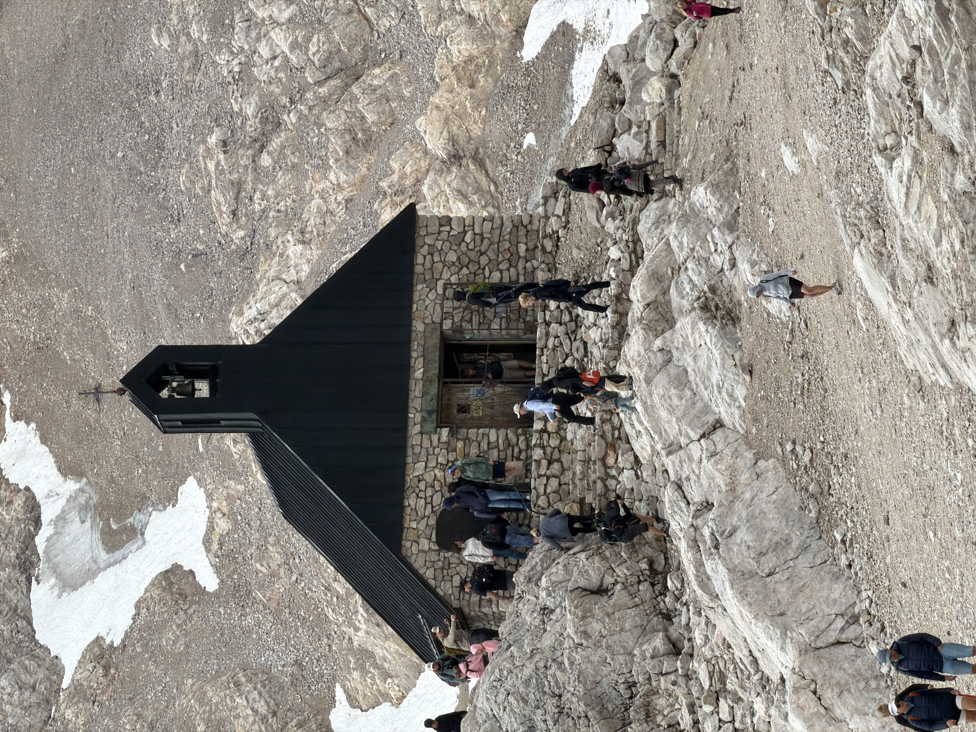 A stone building, possibly a chapel or mountain hut, with a dark, sloped roof stands on a rocky slope. A cross tops the structure's bell tower. Several individuals, appearing to be hikers, are scattered around the building and on the surrounding rocky terrain. Some are ascending or descending the rocky incline, while others are gathered near the building's entrance. The environment is mountainous and appears to be at a high altitude, with patches of snow visible on the ground and higher slopes. The overall scene depicts a stop or point of interest in a mountainous region, likely a popular hiking destination in Garmisch-Partenkirchen, Germany, given the context.