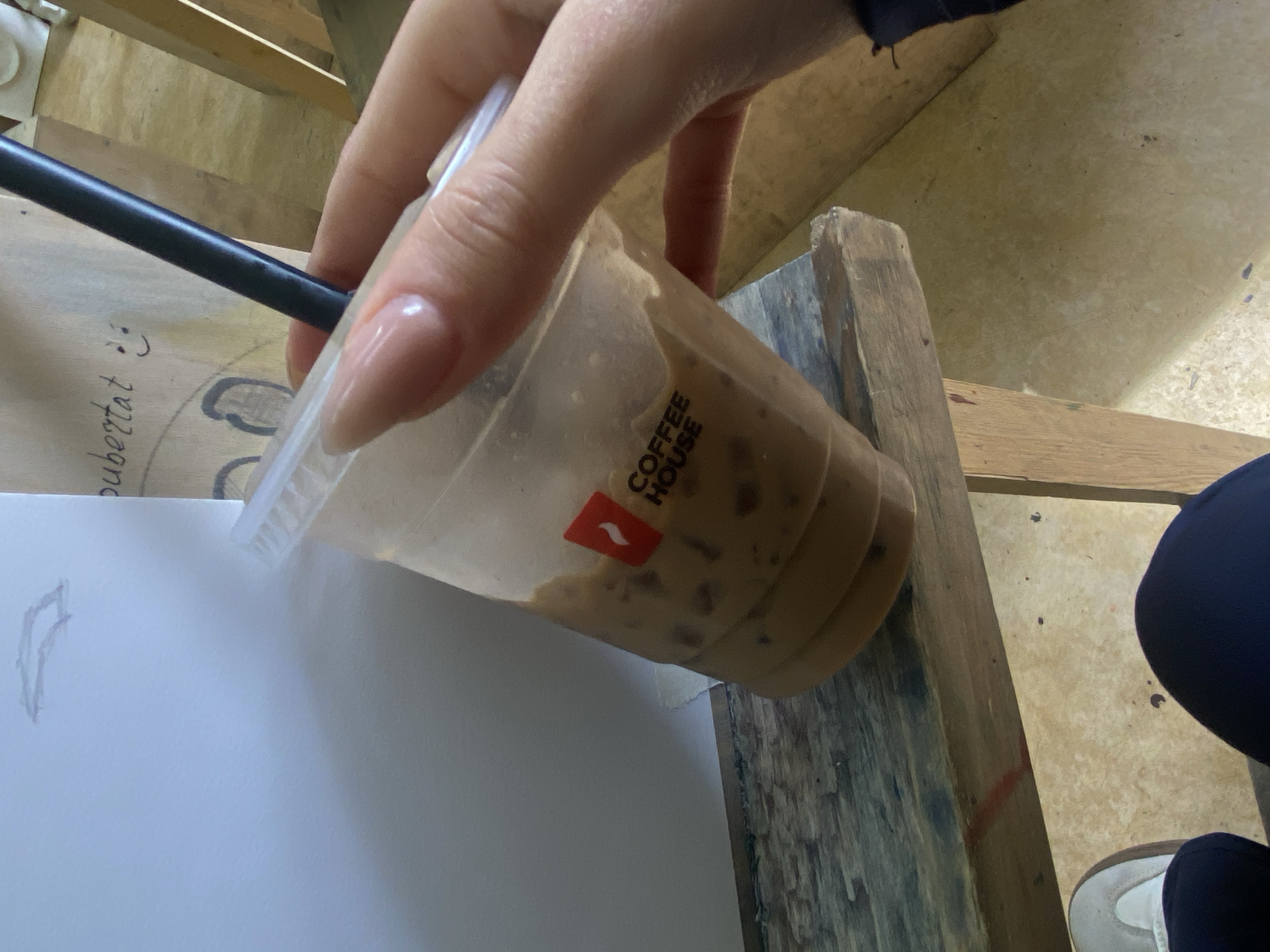 A hand holds a plastic cup containing an iced coffee beverage from Coffee House, with a black straw inserted. The cup rests partially on a wooden surface. A piece of white paper with a drawing and the word "ubertat" with a smiley face is visible. The person is wearing dark-colored pants and a white shoe. The location is possibly Yerevan, Armenia, based on the coffee brand.