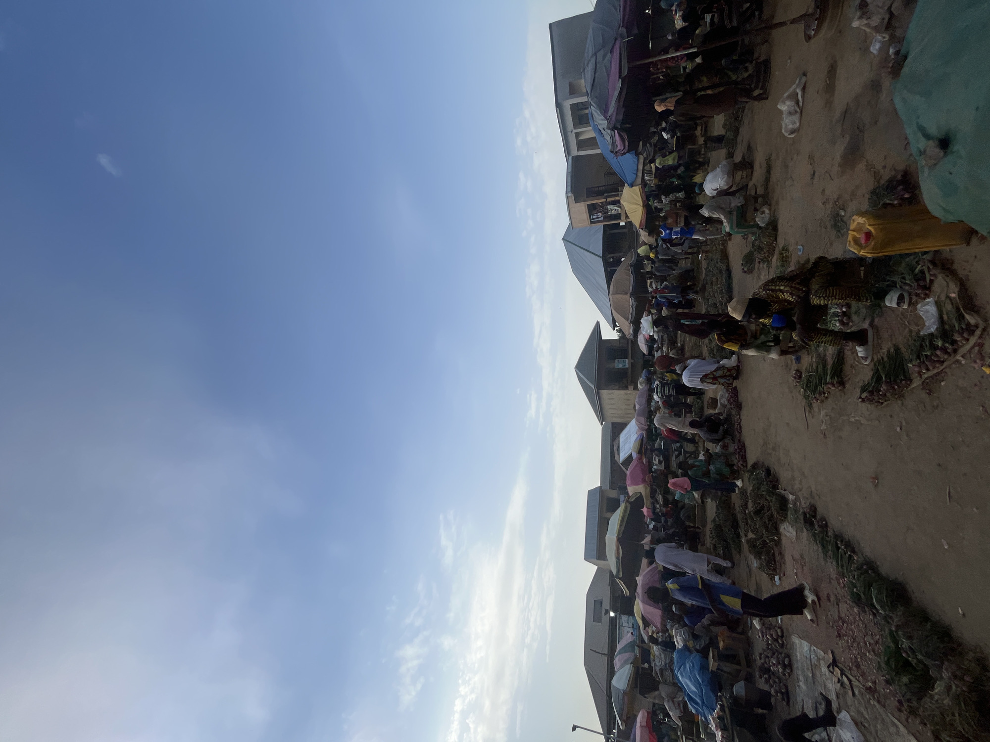 The image depicts a bustling outdoor market scene in Rafin Par, Nigeria. The market is filled with numerous people, many of whom appear to be vendors and shoppers. Stalls are set up with various goods, predominantly what appears to be produce like onions, displayed on the ground and under umbrellas or makeshift canopies for shade. The ground surface is a mixture of dirt and some scattered fabric or tarps. Several individuals are seen sitting or standing, engaged in transactions or attending to their merchandise. The architecture in the background suggests a semi-urban environment with buildings that have pitched roofs. The sky above is clear with some wispy clouds, indicating daylight conditions. The overall impression is one of lively commercial activity in a local marketplace.