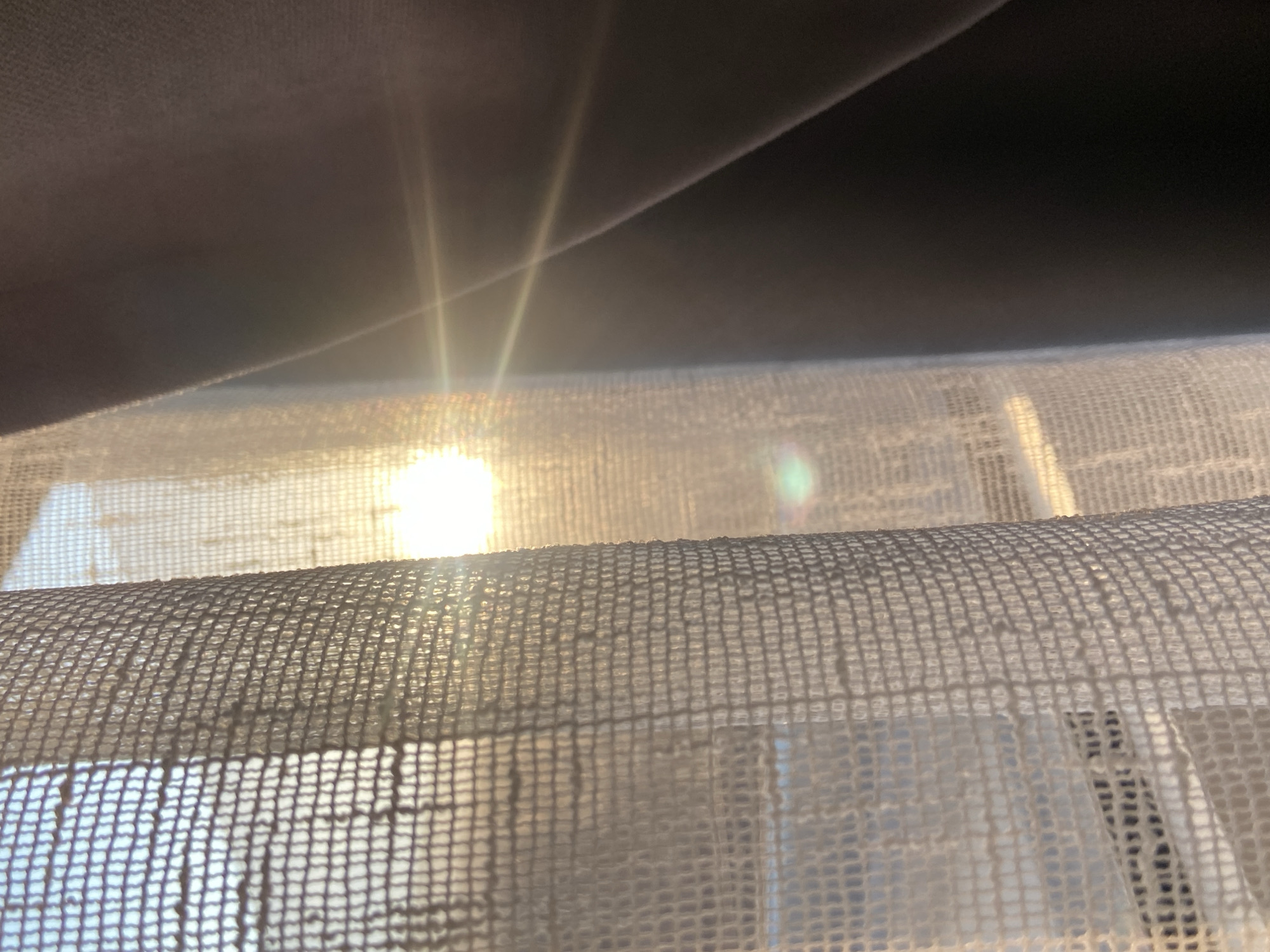 The image depicts sunlight filtering through a white, mesh-like curtain, with a dark fabric draped above. The bright light source is positioned behind the curtain, creating a lens flare effect. The specific location in Budva, Montenegro cannot be determined from this image.