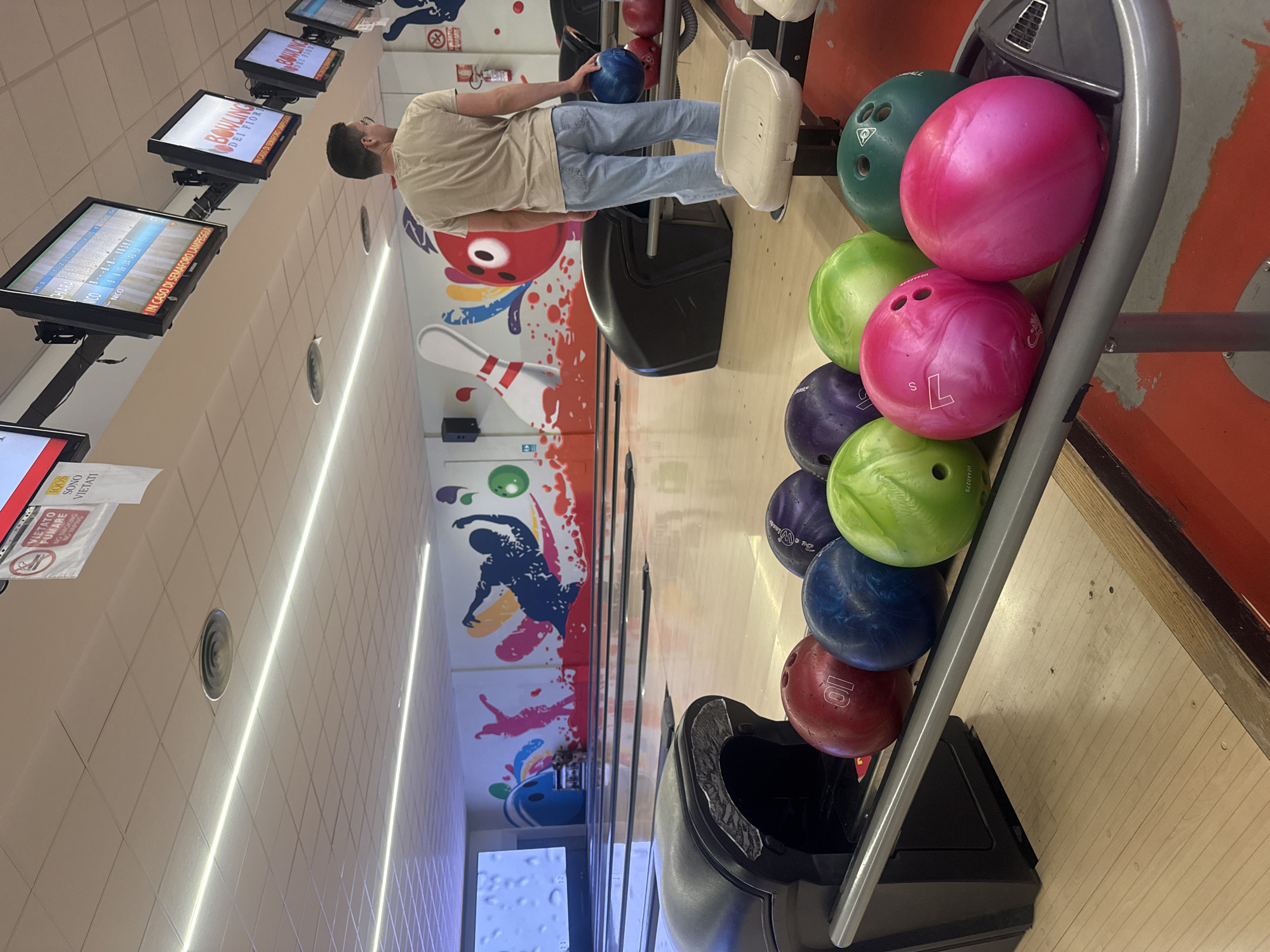 Here is a forensic description of the image:
The image is taken at a bowling alley, in the city of Milan, Italy. A male is seen holding a blue bowling ball, with a rack of bowling balls nearby. The bowling balls are various colors and weights. The lanes are numbered and there are monitors above the lanes displaying scores. The wall behind the lanes has a colorful mural depicting bowling. The lighting in the bowling alley is bright.