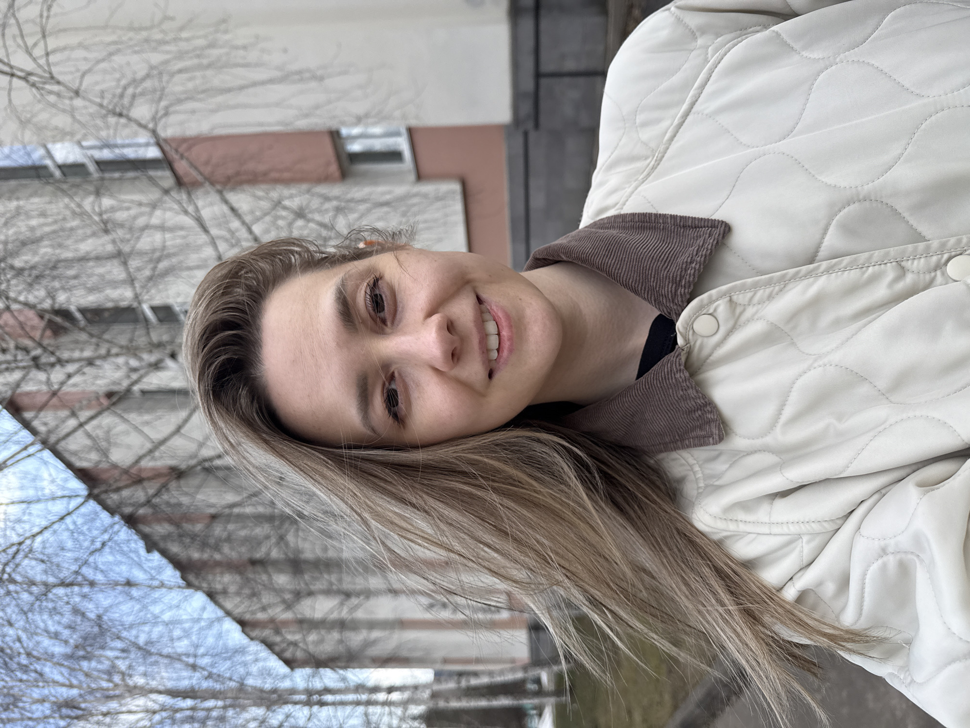 A young woman is depicted in a close-up outdoor portrait, smiling warmly at the camera. She has long, straight, light brown hair that cascades over her shoulders and down her front. Her eyes are dark, and she exhibits a natural, pleasant expression.
She is wearing a light cream or off-white quilted jacket, characterized by a wavy stitch pattern and a contrasting brown corduroy collar. The jacket features light-colored snap buttons visible on the front placket.
The background reveals a typical urban or suburban setting, with several bare, dormant trees suggesting a cooler season like late autumn, winter, or early spring. Behind the trees, light-colored residential buildings, possibly apartment blocks with some dark window or balcony details, are visible. The lighting is diffused, indicating an overcast day. The scene reflects a casual moment in Chaikovsky, Russia. No visible text is present in the image.