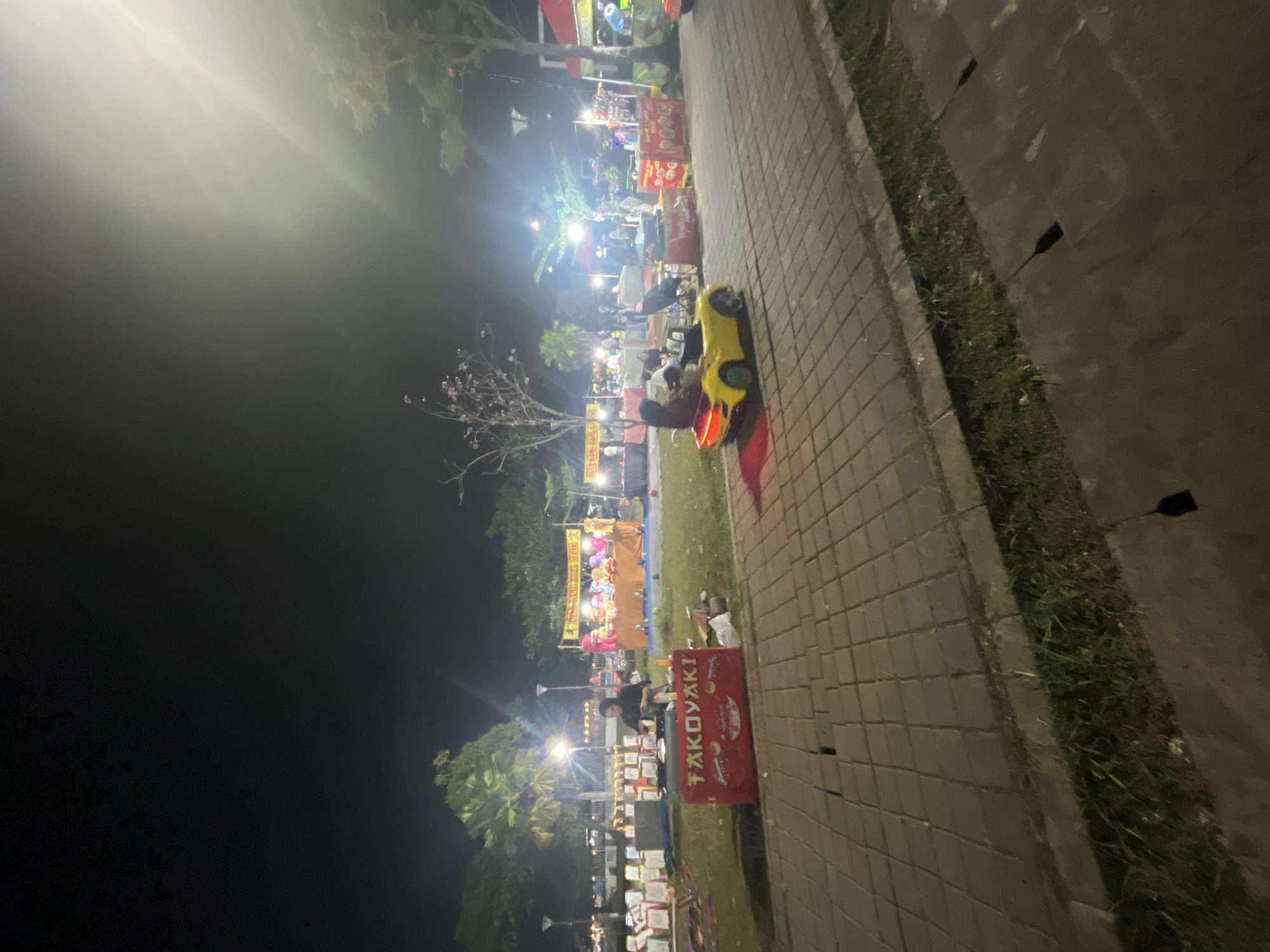 An exterior nighttime street scene in Sodong, Indonesia, captures an open-air food and entertainment market. In the foreground, a paved walkway is lined with grass and drainage grates. To the right, a bright yellow toy car with two occupants, a child and an adult, is illuminated by the scene's ambient lighting. The car is moving along the walkway, heading towards the right of the frame.
Beyond the walkway, a grassy area is populated with various stalls and attractions. A prominent red stall in the middle foreground displays the word "TAKOYAKI" in white lettering and appears to be a food vendor. To its left, a person is attending to a cooking station. Further back, several other stalls are visible, some with colorful banners and what appear to be games or merchandise displays. In the background, trees and streetlights dot the landscape, contributing to the overall illumination of the market. The sky is dark, indicating it is nighttime.