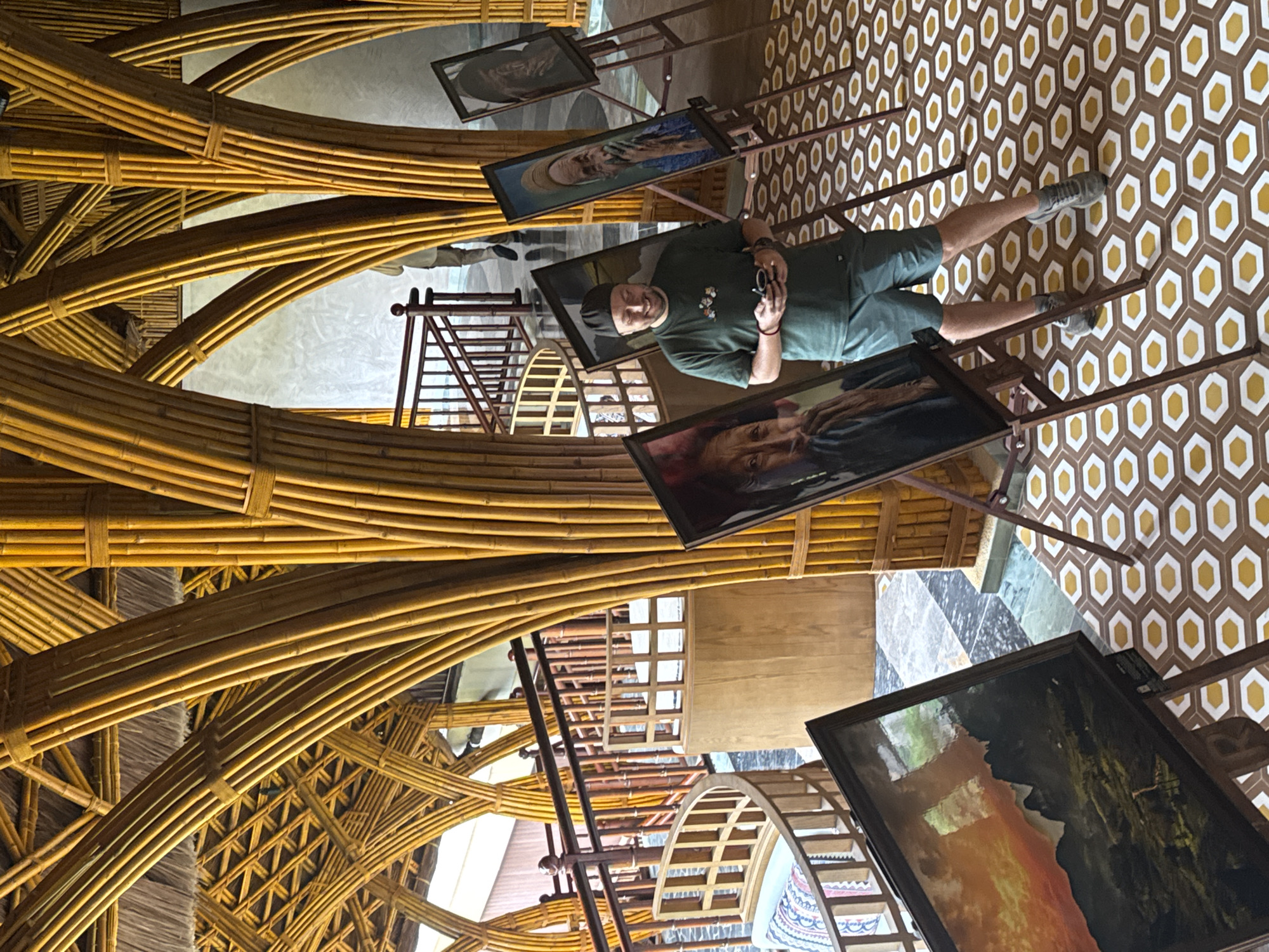 This image depicts an indoor art exhibition or gallery in Púng Luông, Vietnam, characterized by its distinctive architecture. The primary subject is a man standing amidst several artworks, facing the viewer with a slight smile. He is dressed in a dark green t-shirt, dark shorts, a black beanie, and grey sneakers, holding a camera, suggesting he is viewing or photographing the exhibition.
Numerous framed artworks are displayed on wooden easels around the man. Many of these appear to be portraits of older individuals, with one particularly prominent portrait directly to the man's right. Another painting in the bottom right corner showcases a dramatic landscape, featuring a vivid orange-yellow sky that could be a sunrise or sunset, contrasting with dark mountainous or cliff formations.
The setting is notable for its intricate and organic design. The ceiling or structural elements are constructed from numerous bundled bamboo poles, forming elegant arches and interwoven patterns, giving the space a unique, natural, and perhaps traditional aesthetic. The floor beneath has a distinct geometric pattern of brown tiles interspersed with white and yellow hexagons. In the background, elements of wooden stairs or railings and further sections of the building are visible. The overall scene suggests a cultural visit, with the man engaging with the art in a thoughtfully designed environment. No discernible text is visible on the artworks or within the scene.