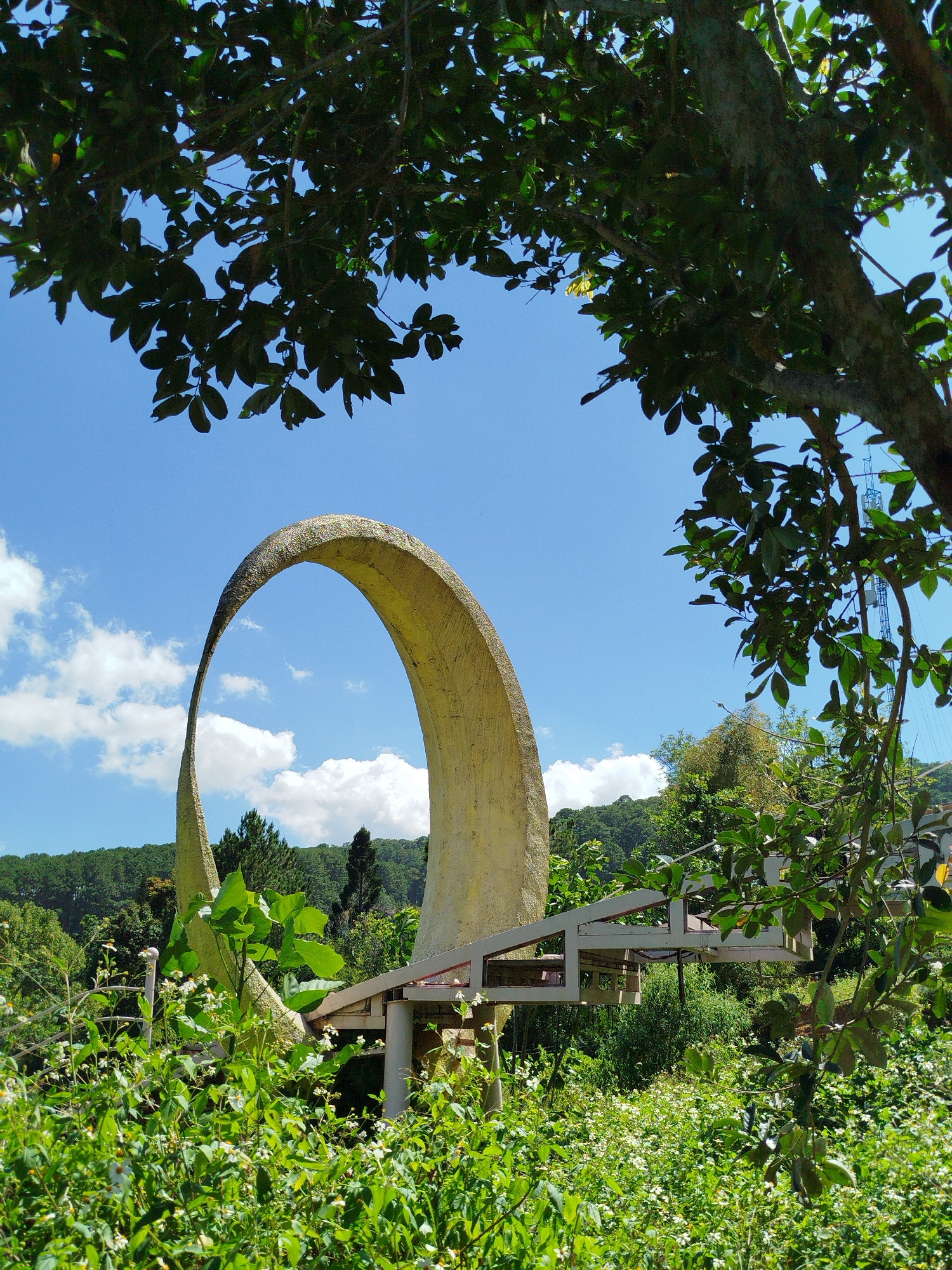 A large, pale yellow, curved concrete structure resembling a large loop or arch stands prominently in a lush, green outdoor setting. The structure is supported by a metal framework, and a dense field of wildflowers and foliage surrounds its base. In the background, a hillside covered with pine trees rises against a bright blue sky dotted with fluffy white clouds. The overall scene suggests a park or garden area. The architectural element is located in Da Lat, Vietnam, a city known for its French colonial architecture, pine forests, and gardens.