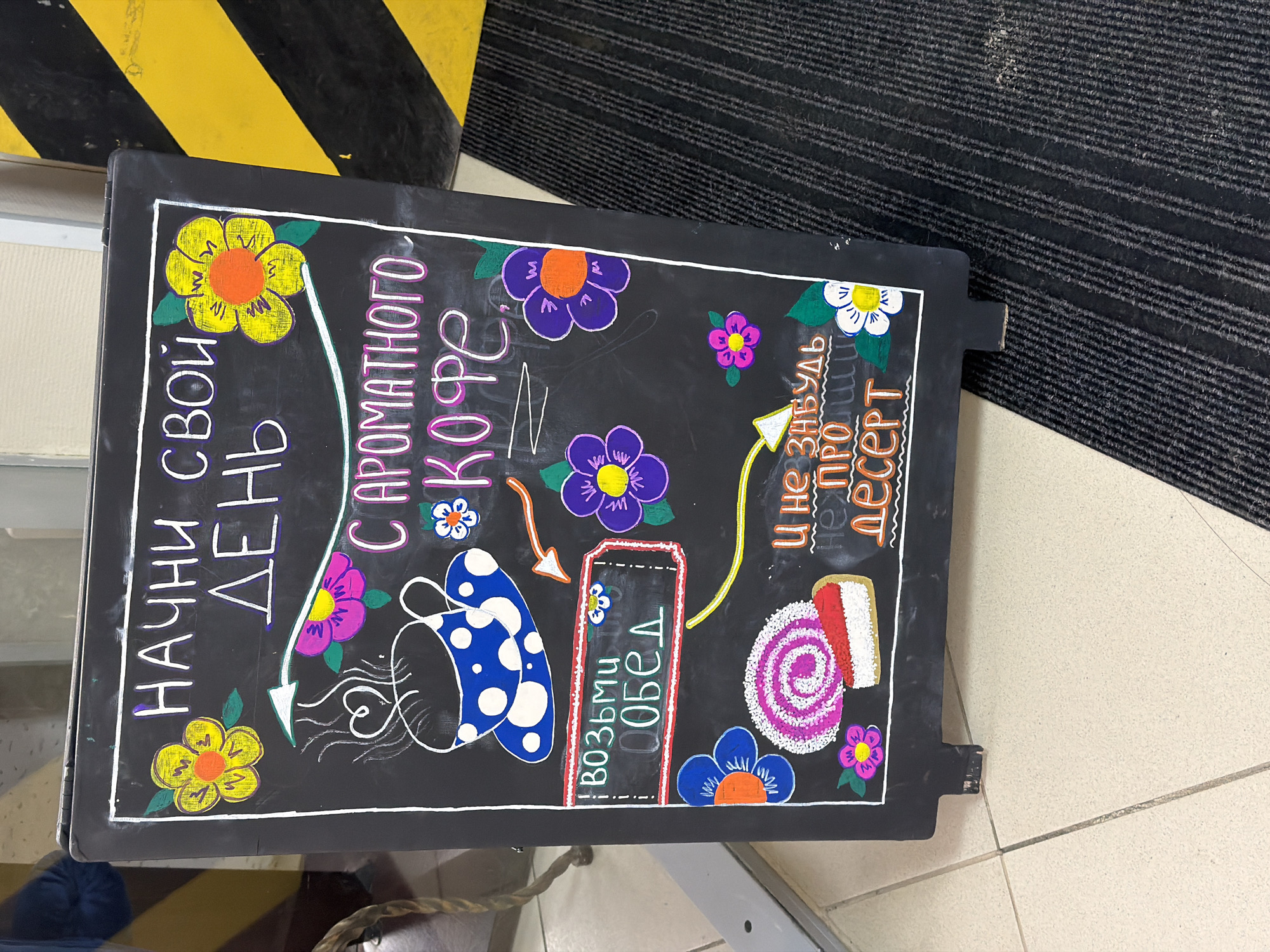 The image shows a black chalkboard sign, likely situated indoors in Berezniki, Russia, displaying colorful hand-drawn lettering and illustrations. The text encourages passersby to start their day with aromatic coffee and to grab lunch, while also reminding them not to forget dessert. The drawings include flowers, a polka-dot cup of coffee, and a sliced cake roll. The sign is propped against a yellow and black striped edge or wall, and a gray striped mat or carpet is visible in the background. The floor is tiled in a light color.