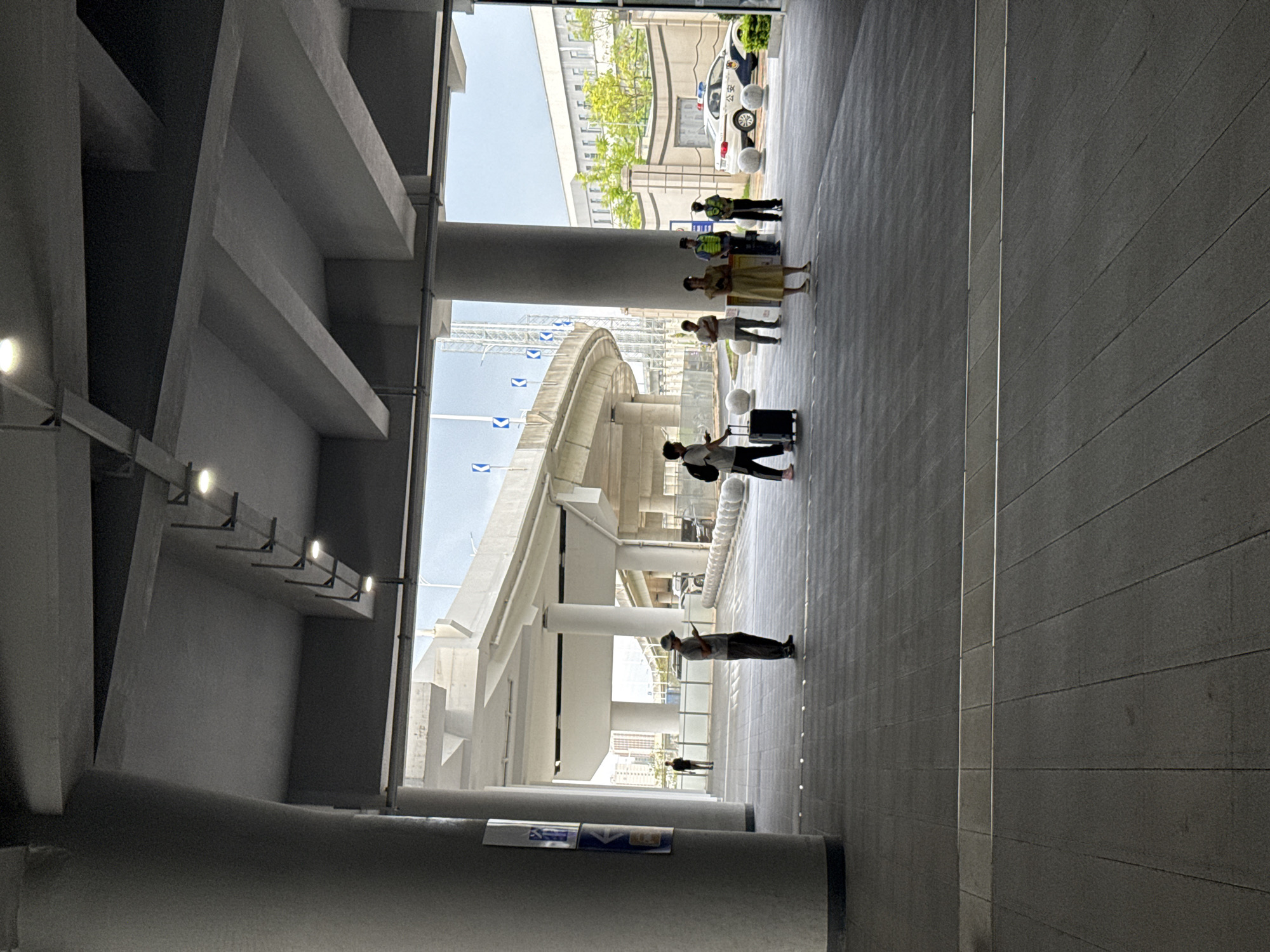 The image depicts a group of people standing and walking in an outdoor, covered walkway, likely part of a public transportation hub or a modern building complex in Huizhou, China. The scene is characterized by strong architectural elements, including concrete pillars, overhead structures with lighting, and a textured gray tiled floor. Sunlight streams in from an opening to the right, suggesting it's daytime.
Several individuals are visible. In the background, near a police car, stand three people in uniform, possibly security or law enforcement officers. In the middle ground, a group of people are gathered. One woman in a light-colored dress stands with her arms at her sides. Two men are standing near her, and one is pulling a rolling suitcase. Further to the right, a person in a light-colored shirt and dark pants is standing and looking at their phone, also with a suitcase nearby. In the far distance, two figures are walking away.
The architecture suggests a contemporary, possibly elevated, structure with curved elements, hinting at a transit line or a bridge. The presence of the police car and uniformed personnel could indicate a security checkpoint or a public area with ongoing operations. The overall impression is one of organized public space, with people engaged in travel or waiting. The lighting suggests it is daytime, and the sky visible through the openings appears clear. There is no visible text in the image that provides specific information about the location or event.
