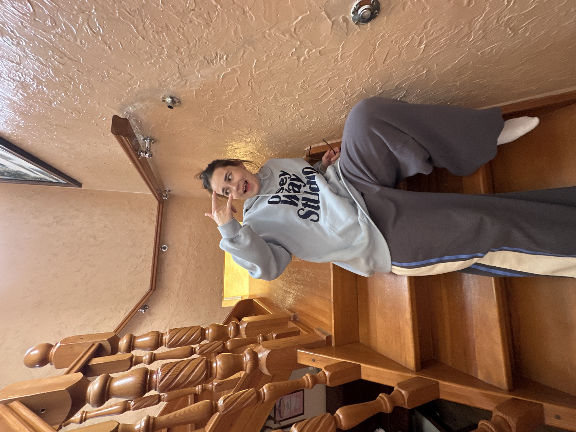 A young woman poses on a wooden staircase. She wears a light blue hooded sweatshirt with the text "Disney World Studios" in navy blue letters. She is also wearing gray sweatpants and a white sock is visible on her right foot. She is looking at the camera and making a hand gesture with her left hand. The background includes a textured, light orange ceiling with light fixtures, a wooden railing, and a framed picture on the wall. There is not enough information in the image to suggest that it was taken in Ilyinsky, Russia.