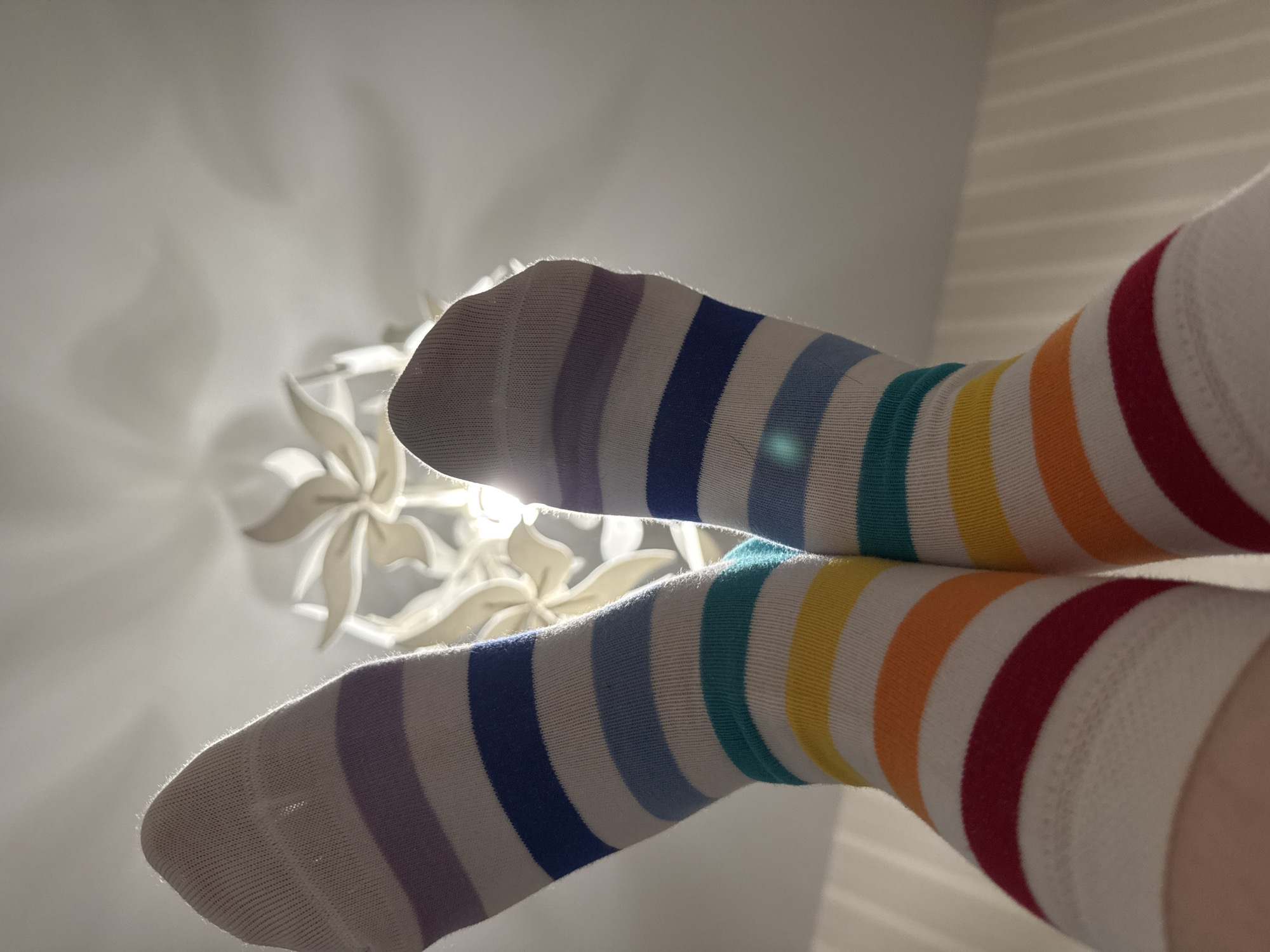 The image captures a low-angle perspective looking upwards, featuring two feet adorned in vibrant rainbow-striped socks. The feet are positioned in the foreground, slightly angled upwards and apart. The socks display horizontal stripes in a full spectrum of colors, including purple, blue, green, yellow, orange, and red, on a white background. They appear to be knee-high or thigh-high.
In the background, a decorative ceiling light fixture or fan is visible. It is white or light-colored, designed with several petal-like or abstract floral elements. A strong light source, possibly sunlight from a window or the fixture itself, is positioned behind the feet, creating a bright halo effect around their outlines and illuminating fine fibers on the socks. This light also casts soft shadows on the pale ceiling and walls. On the right side of the frame, distinct linear shadows suggest light entering through blinds or a window.
The scene depicts a moment of relaxation, with someone lying down and their feet elevated. The bright, natural light suggests it is daytime. The overall atmosphere is light, cheerful, and comfortable, amplified by the colorful socks. There is no visible text in the image. The location context is Chaikovsky, Russia, within an indoor setting.