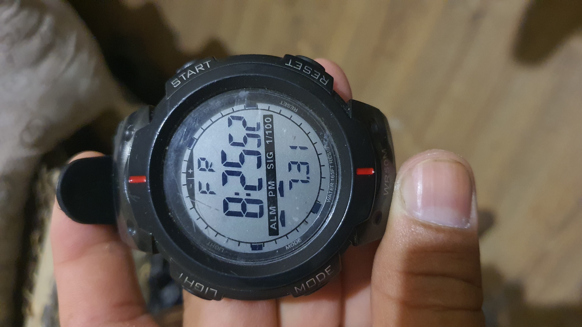 A close-up shot shows a digital watch face with black casing and a grey dial. The watch is held by a person's thumb and forefinger, with the thumb positioned at the top of the watch and the forefinger near the bottom. The watch face displays the time as 8:25:52, with "FR" above it, likely indicating Friday. Below the time, "ALM PM SIG 1/100" is visible, suggesting alarm, PM, signal, and a stopwatch function. The number "7.31" is displayed below that. The watch also has labels around the dial: "LIGHT" on the left, "START" on the top right, "RESET" on the bottom right, and "MODE" on the bottom left. The watch is also labeled "WATER 165FT RESIS" and "WR50M" indicating water resistance. The background is blurred, appearing to be a textured surface, possibly a couch or fabric, in muted tones. The scene is not identifiable as being in Karankul, Uzbekistan.