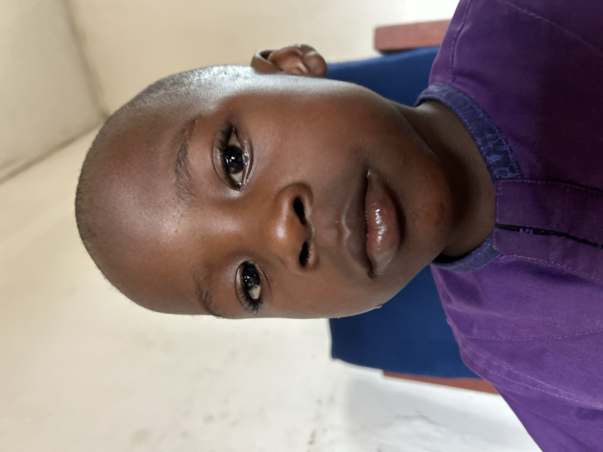 A medium close-up shot depicts a young, Black child with a closely shaved head. Their skin has a smooth texture. The child is wearing a purple shirt. The background appears to be an indoor setting with white walls and a blue object visible behind them.
