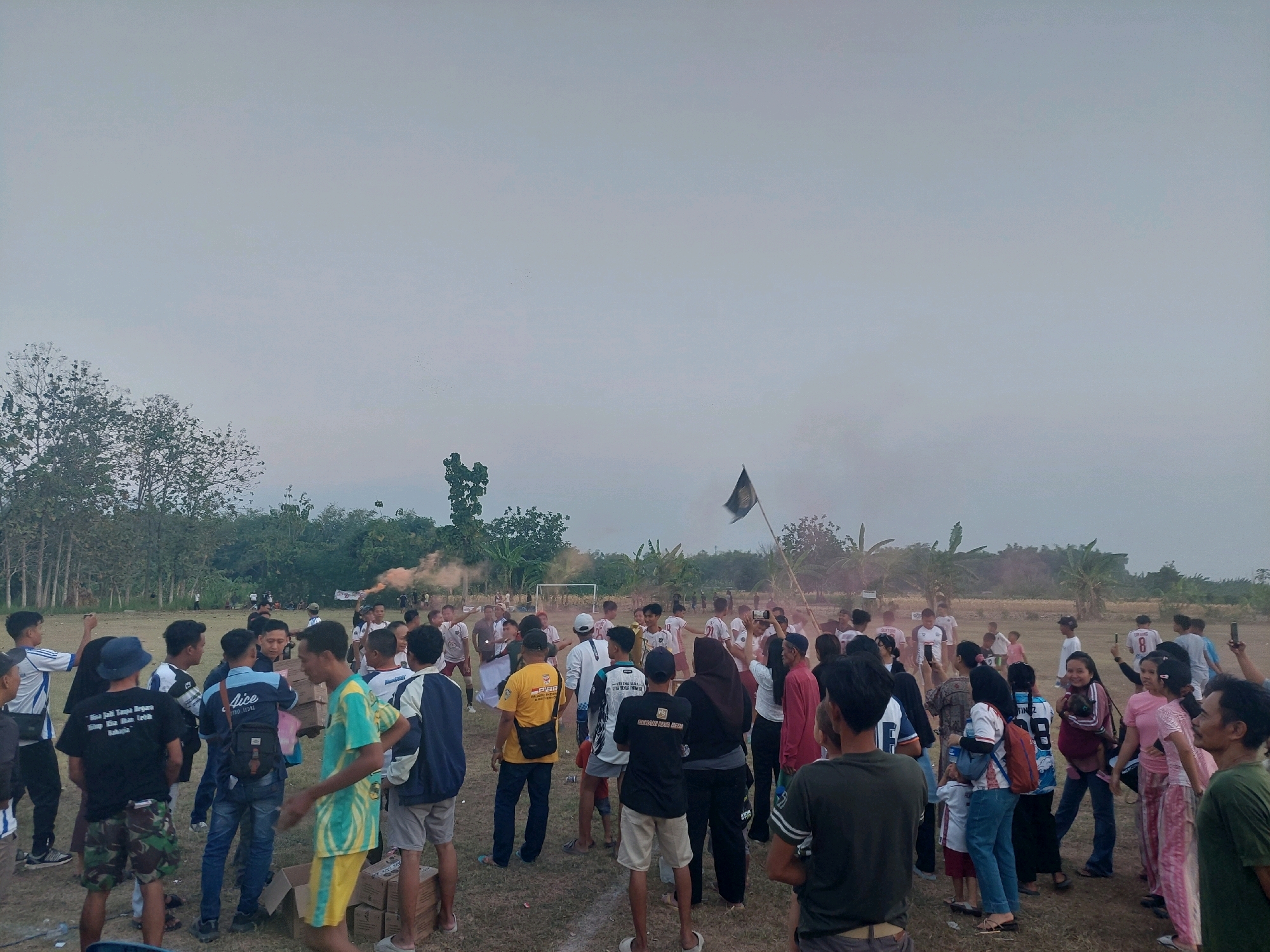 The image depicts a gathering of people in an outdoor grassy area, possibly a field or park, with trees and vegetation in the background. The sky is overcast and a pale grey. A group of people, predominantly young men and some women, are standing and interacting in the foreground and mid-ground. Many are wearing casual clothing, including t-shirts, shorts, and pants. Some are holding boxes or carrying bags. In the mid-ground, a few individuals are holding up what appear to be flares or smoke bombs, emitting pink and orange smoke. A flag on a pole is visible in the distance, and a soccer goal is also discernible. Several people are looking towards the smoke. The environment suggests a communal event or celebration taking place outdoors. The location is identified as Ngablak, Indonesia.