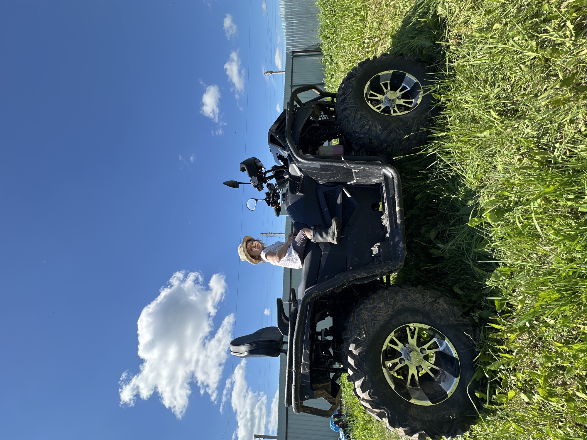 In Kharnavy, Russia, a young child wearing a straw hat, a white t-shirt, and dark shorts sits on a black all-terrain vehicle (ATV) parked in a grassy field under a bright blue sky with scattered white clouds. The ATV has large, knobby tires with silver rims and a black passenger seat. A metal fence runs along the horizon behind the vehicle, with a utility pole visible in the distance. The ground is covered in lush green grass, with some areas appearing recently cut.