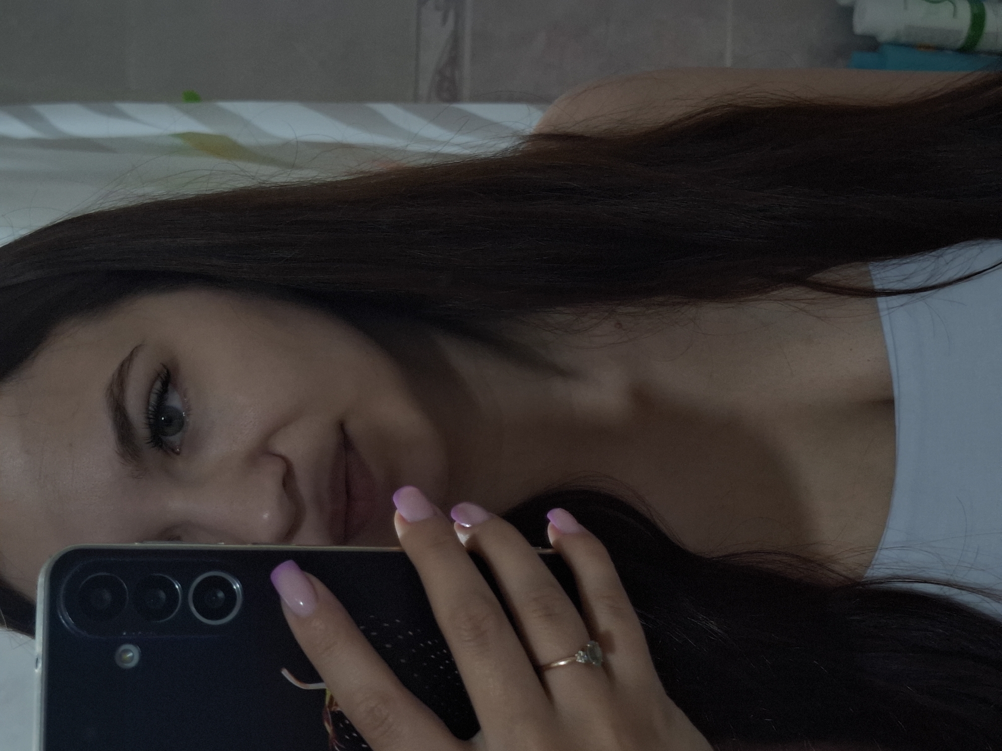 A woman is taking a selfie indoors. She has long, dark brown hair and light skin. She is wearing a light-colored top. She is holding a dark-colored smartphone with a ring on her finger and nails that are painted with pink tips. The background appears to be a bathroom with a tiled wall and toiletries visible in the upper right corner. The location is not discernable as Chaikovsky, Russia.