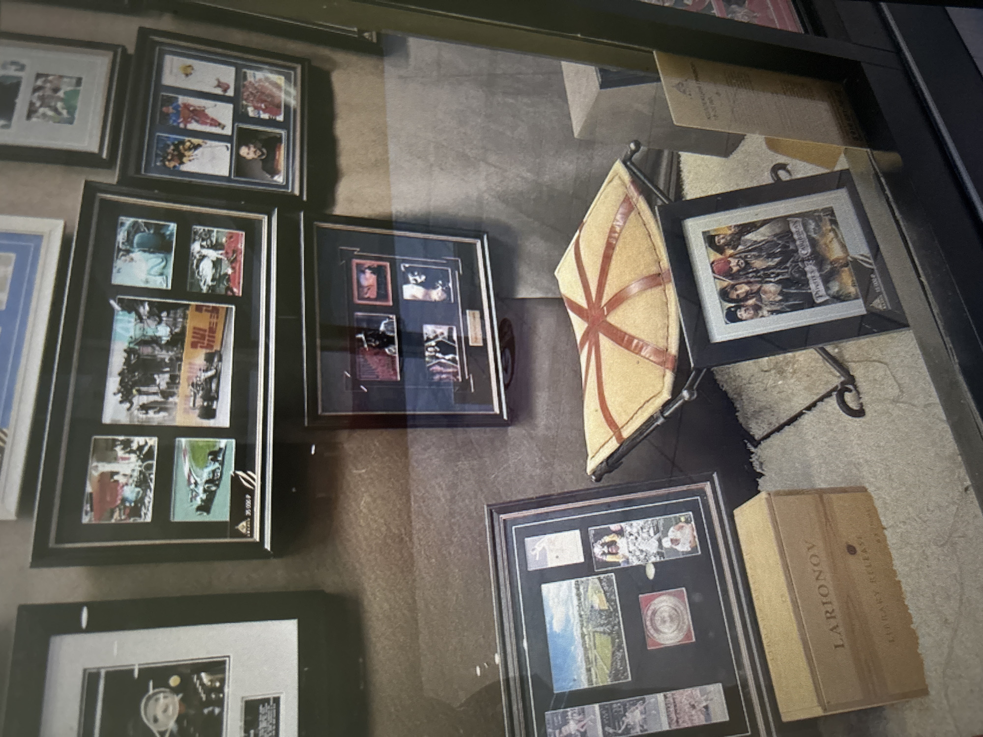 The image shows a display case with several framed items and a wooden box. The display case appears to be a retail or exhibit setting. Among the framed items are photographs related to sports, possibly racing and hockey, and a framed movie poster from "Pirates of the Caribbean." There is also a small stool-like object with a yellow cushion. A wooden box with the text "LARIONOV" and "LIBRARY RELEASE" is visible on the floor. The overall impression is one of memorabilia display, possibly in a public venue. There are no people visible in the scene.