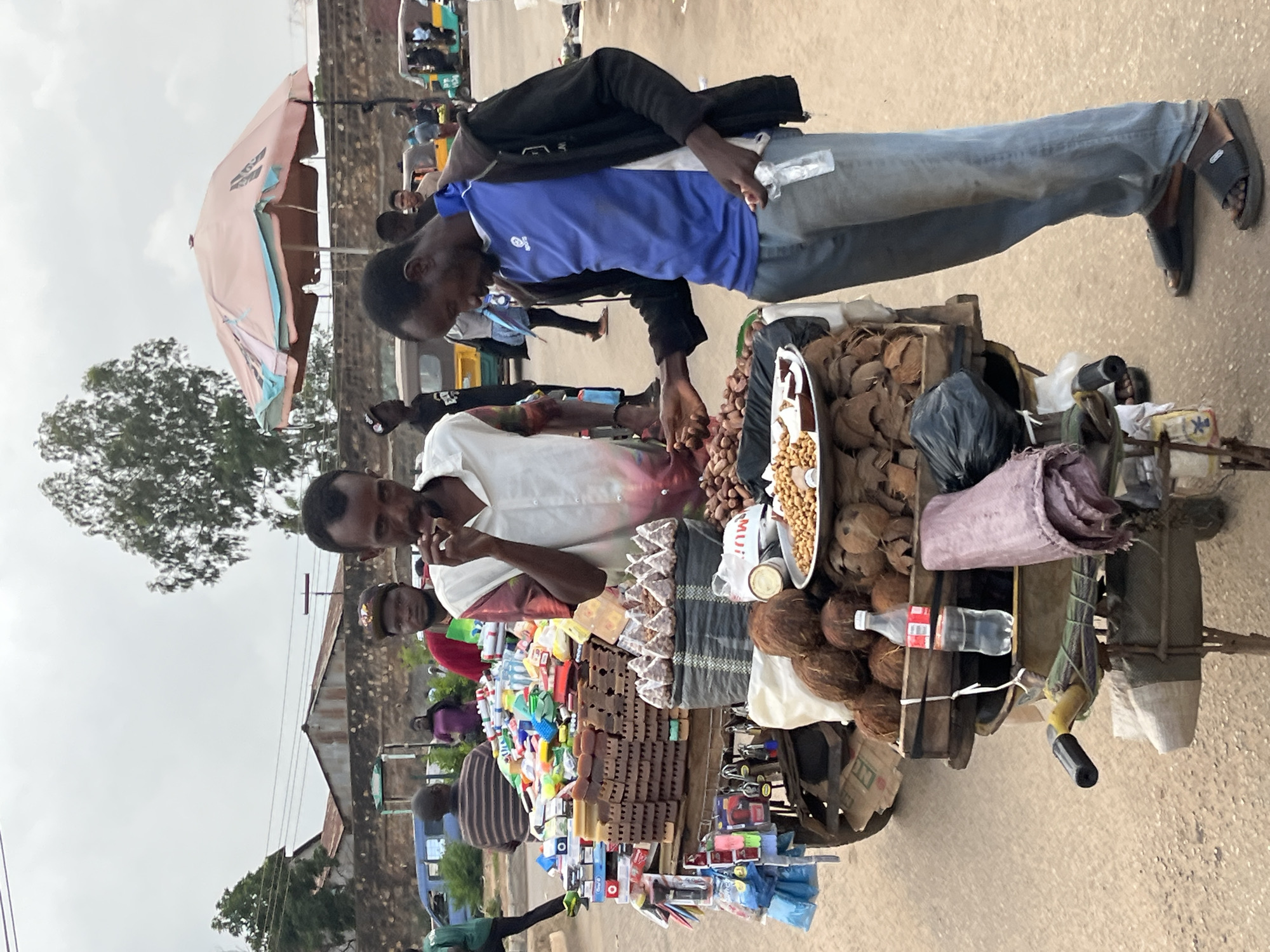 A street vendor in Bukuru, Nigeria, stands behind a cart laden with goods. The cart is packed with an assortment of items, including coconuts, nuts, packaged snacks, and various small consumer products such as locks and cleaning supplies. Two men are actively engaged with the cart. One man, wearing a blue shirt and black jacket, is selecting nuts from a tray. The other man, in a white shirt, appears to be interacting with the same vendor or observing. In the background, other individuals and vehicles, including what appear to be auto-rickshaws, are visible, suggesting a busy market or street environment. The sky is overcast, and the ground is unpaved.