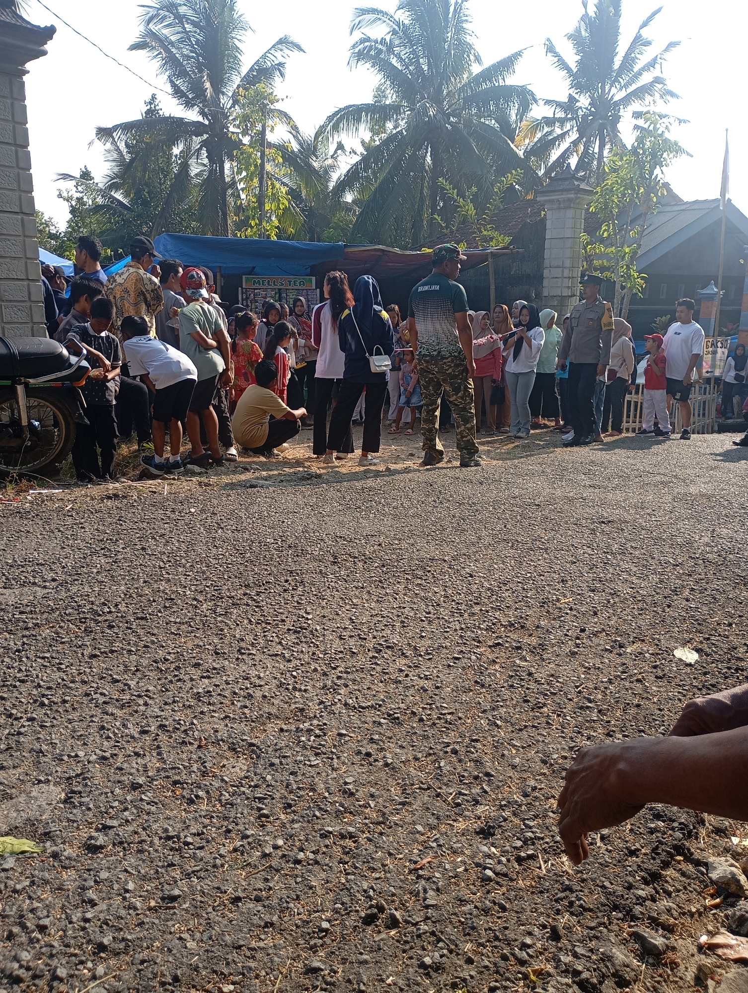 The image depicts an outdoor scene, possibly in Ketepung, Indonesia, with a gathering of people. The ground is covered with gravel and scattered dry grass. In the background, palm trees and houses are visible under a bright sky. A blue tarp is set up, with a sign that reads "MELL'S TEA." Various individuals are standing and walking around, including children and adults. Some appear to be in a queue or observing an event. A police officer in uniform is present, suggesting a possible community event or gathering. In the foreground, the hands of an individual are visible, resting on the gravel. A motorcycle is parked on the left side of the image. The overall atmosphere appears to be casual and communal.