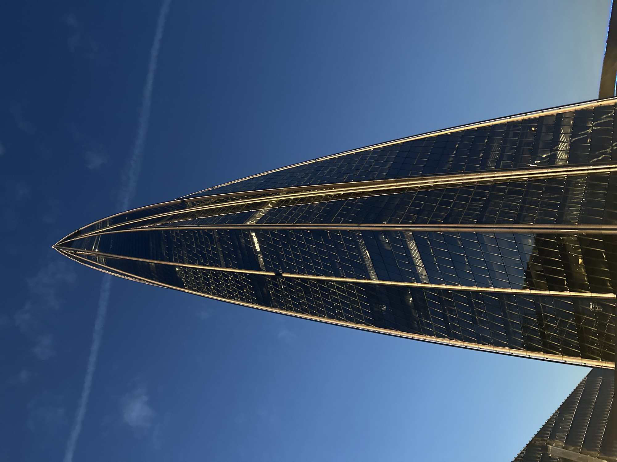The image shows the Lakhta Center skyscraper in Saint Petersburg, Russia, under a clear blue sky with a faint contrail. The skyscraper, with its distinctive pointed, spiraling design, is covered in reflective glass panels and illuminated by exterior lights. The lower left portion of the image also shows a glimpse of another modern building with a similar glass facade. The time of day appears to be either early morning or late evening due to the warm lighting on the skyscraper against the deep blue sky. There are no people or active elements visible in the scene.