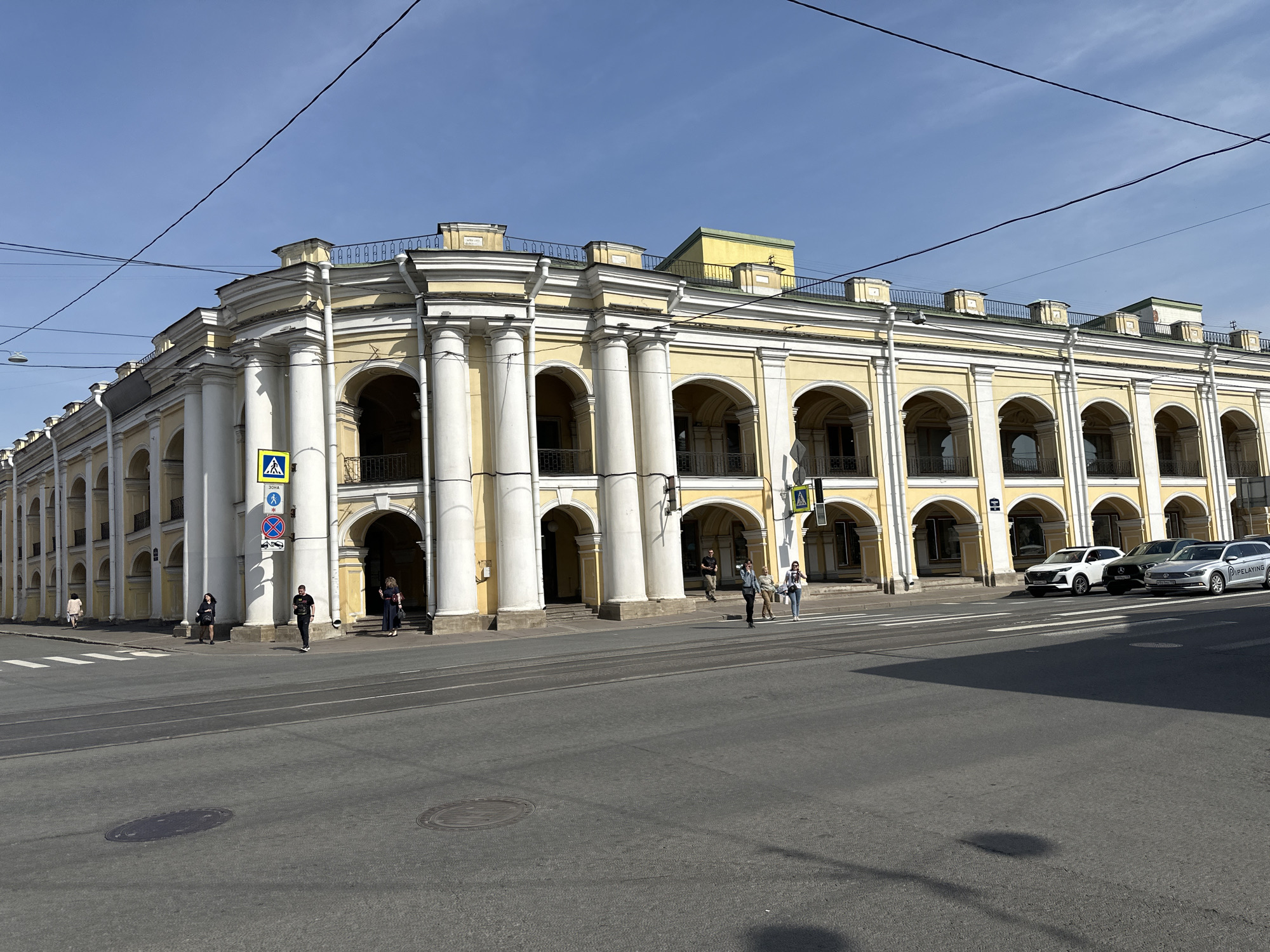 The image depicts a street scene in Saint Petersburg, Russia. The dominant feature is a long, yellow building with a distinctive architectural style characterized by numerous white columns supporting arched openings. The building extends along the street, with a visible rooftop area featuring a railing.
In front of the building, several people are present: some are standing near the building's entrance, while others are crossing the street at a pedestrian crossing marked with a 'zebra' pattern and a pedestrian crossing sign. On the right side of the image, several cars are parked.
The street itself is wide and paved, showing typical urban street markings. The sky is clear and blue, suggesting a sunny day. Power lines are visible overhead, crossing the sky at angles. There are manhole covers in the foreground road, adding to the street's detail.