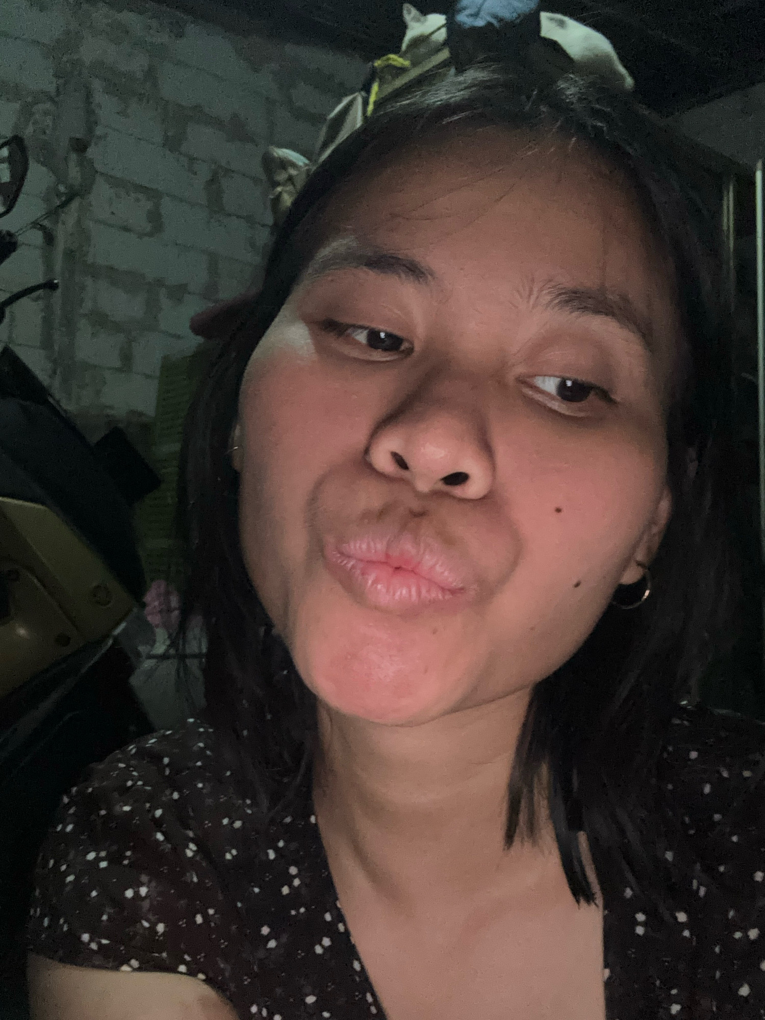 The image is a close-up, selfie-style photograph of a woman with dark hair and brown eyes, puckering her lips. She is wearing a dark top with a white polka dot pattern. The background appears to be an interior setting with a brick wall and what looks like a motorcycle on the left. The lighting is dim, casting shadows. The scene is likely in East Jakarta, Indonesia, based on the provided context, though no specific landmarks or definitive indicators are visible within the frame to confirm the exact location.