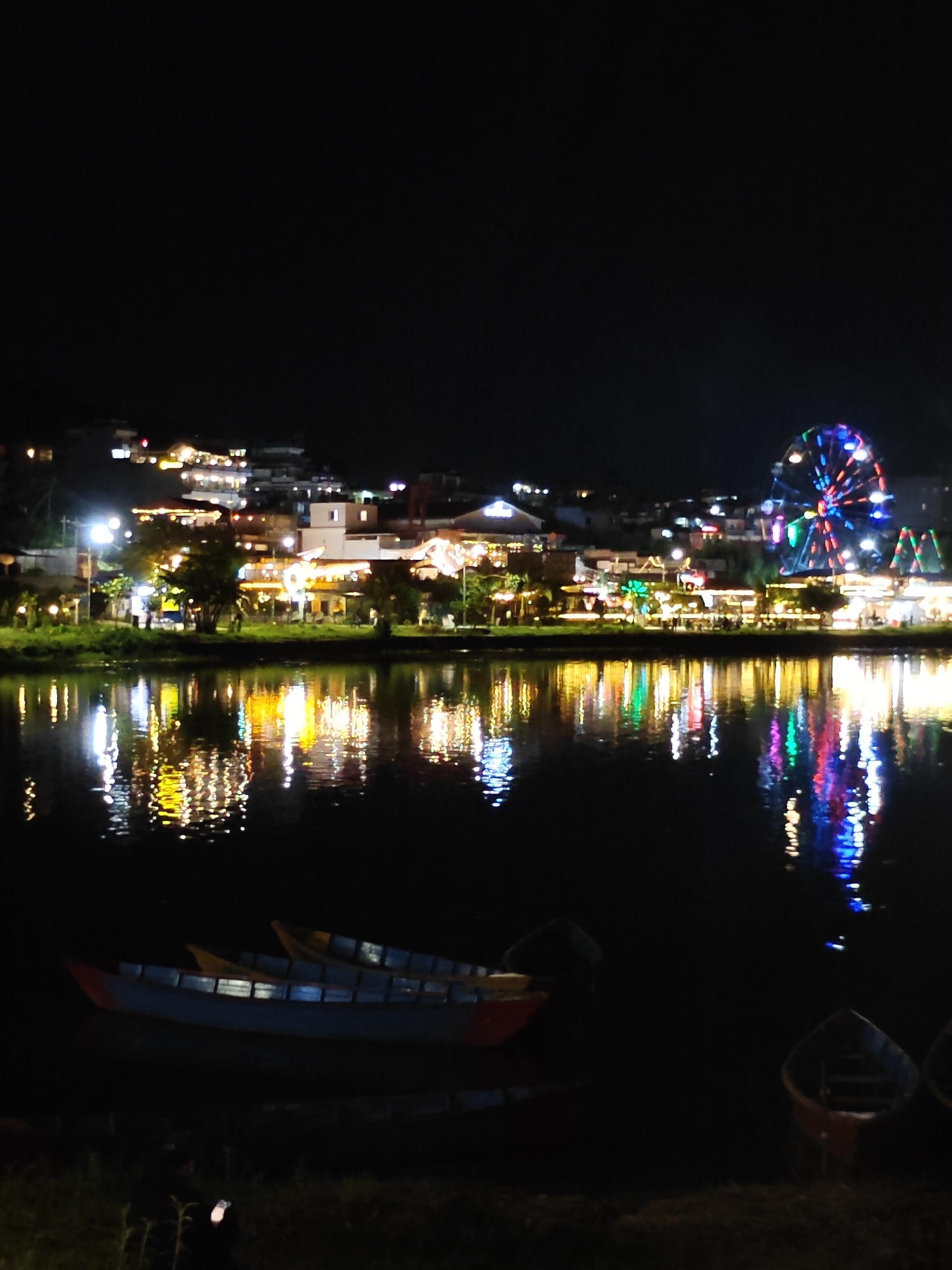 The image depicts a nighttime scene of Phewa Lake in Pokhara, Nepal, with the Lakeside area illuminated by lights. In the foreground, two traditional Nepali boats are moored on the dark water. The middle ground shows the tranquil surface of the lake reflecting the vibrant lights of the buildings and a Ferris wheel situated on the shoreline. The buildings, a mix of shops, restaurants, and hotels, are brightly lit, creating a festive atmosphere. The Ferris wheel, a prominent feature of the Pokhara skyline, is adorned with colorful lights. In the background, the dark silhouette of the surrounding hills is visible against the night sky. The overall impression is one of a bustling tourist destination at night, with the natural beauty of the lake enhanced by the artificial lights.
