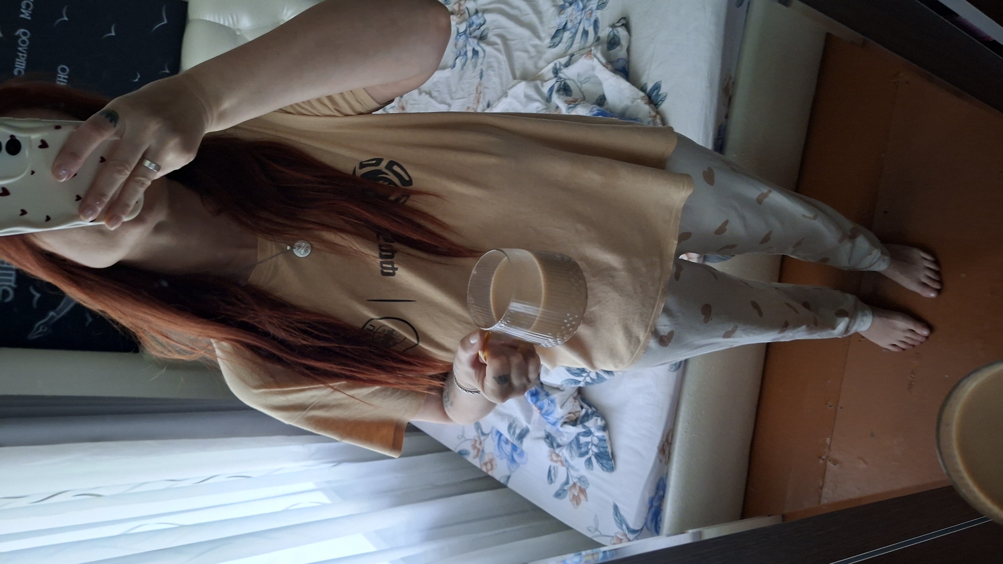 A person with reddish-brown hair is standing in a bedroom in Myadzyel, Belarus, holding a glass of coffee. They are wearing a peach-colored t-shirt and light-colored pajama pants with a heart pattern. The background shows a bed with blue and white floral bedding, sheer white curtains, and a dark wall with artwork. A mirror reflects the scene, showing the person taking the photo with their phone. The floor is made of brown wood.