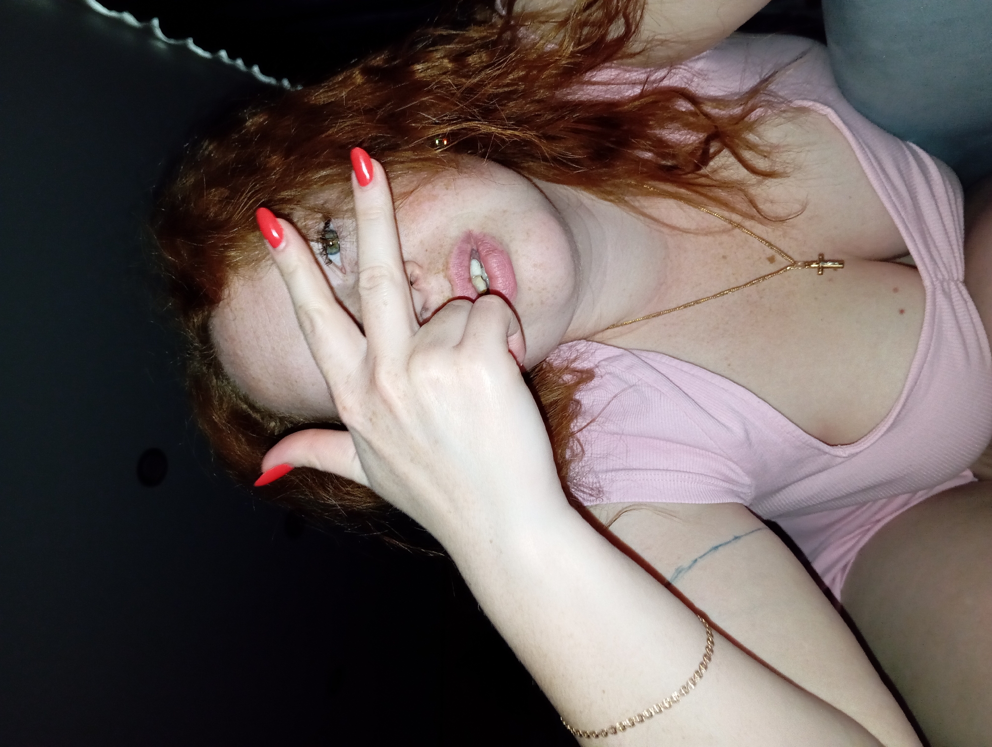 A woman with fair skin and red curly hair is angled in the image. She is wearing a pink v-neck top and a gold necklace with a cross pendant. She has red nail polish on her fingers. Her right hand is raised near her face, with the ring and middle fingers touching her lips. Her left arm, visible at the bottom right, has a gold bracelet and a faint blue line, likely a tattoo. The background is dark, with a white string of lights visible at the top. The location is unspecified and cannot be determined based on the visual information provided.