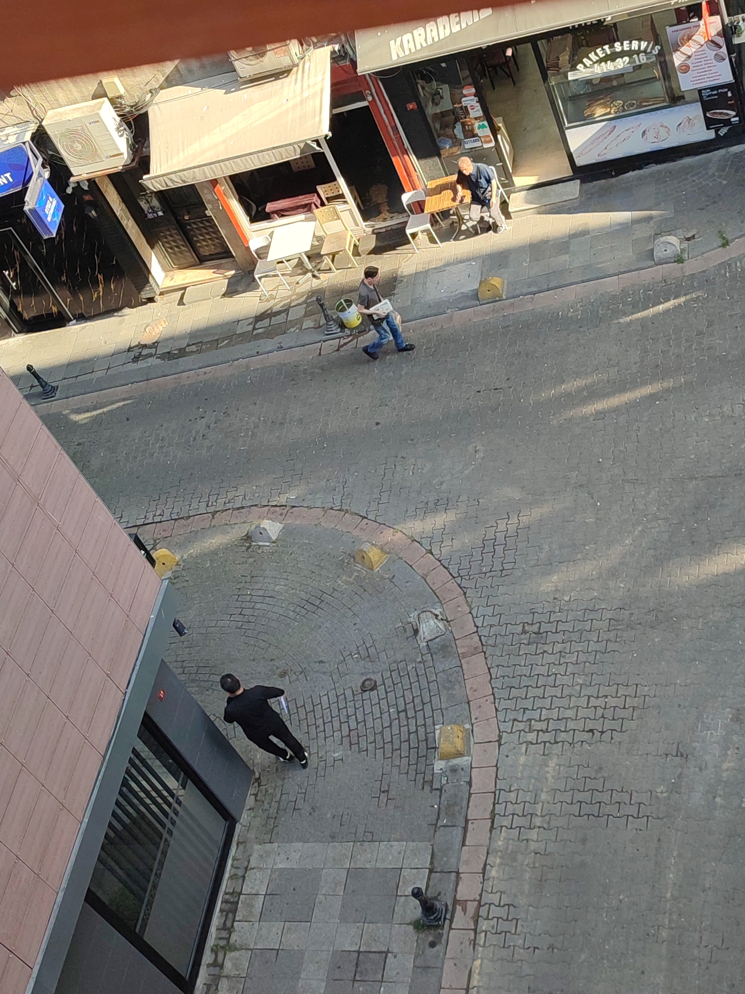 The image shows a street scene from an elevated perspective in Kadıköy, Turkey. In the foreground, a person in a black outfit walks on a paved sidewalk, carrying a white object. To the right, a street with a cobblestone-like pattern is visible, with sunlight casting shadows. Further up the street, a shop with a red awning is prominent. Two individuals are near the shop entrance: one person is seated at a small table outside, and another person is walking away with a bucket and a tool. Another shopfront, featuring a display case with food items and advertisements, is also visible. The environment includes building facades, shop signs, outdoor seating, and street furniture.