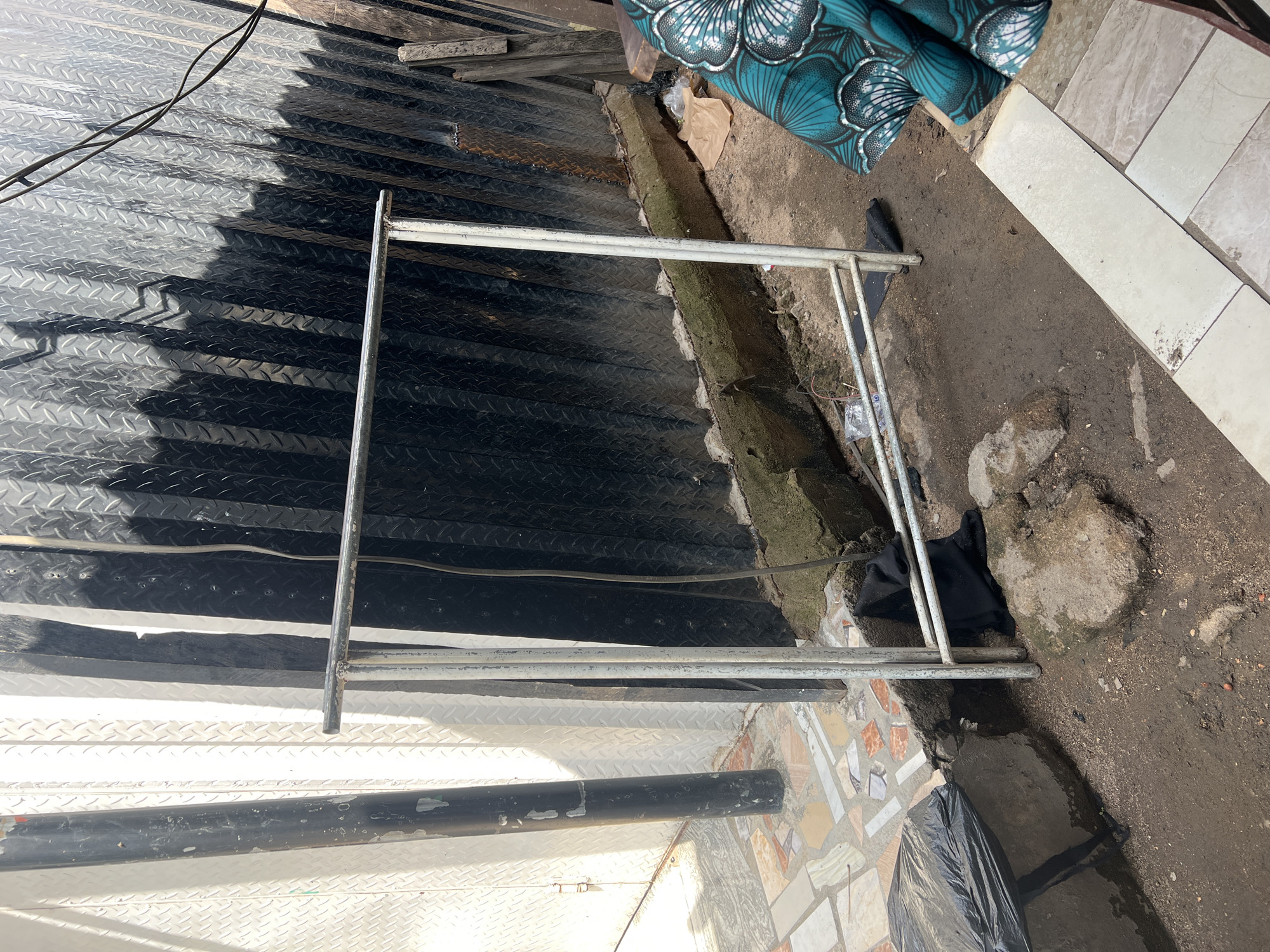 The image depicts an outdoor scene in Jalingo, Nigeria. In the foreground, a white metal clothing rack stands on a patch of dirt and scattered stones. A black piece of fabric is draped over the lower bar of the rack. To the left, a large black plastic bag rests on a concrete block. Behind the rack, a wall made of black corrugated metal is visible, with a portion of a white corrugated metal wall to its left. A black cylindrical pole stands against the white wall. The ground is a mix of dirt, scattered tiles, and concrete. In the upper right corner, a piece of fabric with a teal and white floral pattern is visible. Electrical wires are strung across the top of the corrugated metal wall. The overall impression is one of a utilitarian outdoor space, possibly a backyard or workshop area.