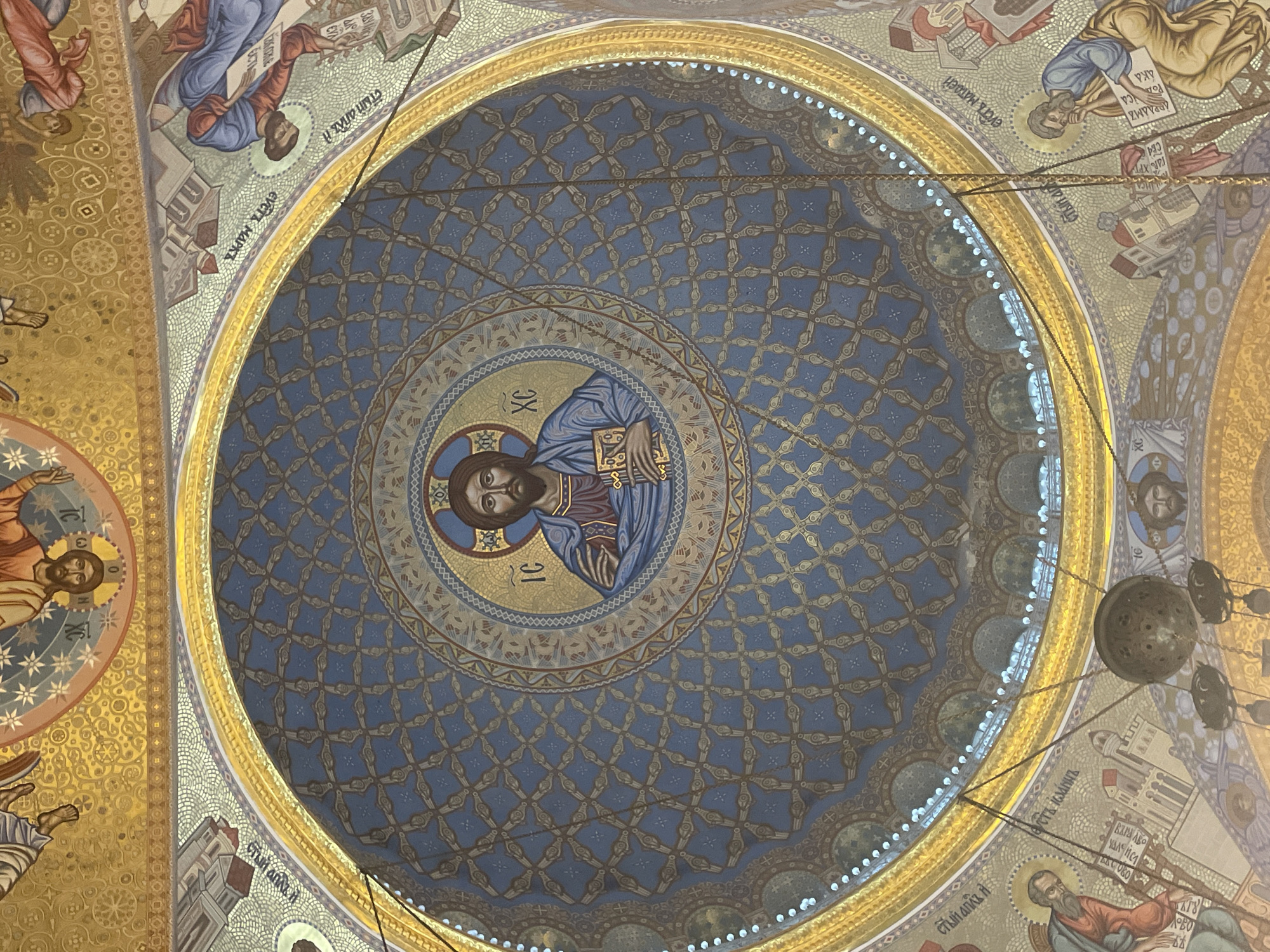 The image shows the interior of a domed ceiling decorated with religious frescoes and mosaics. In the center of the dome, a mosaic depicts Jesus Christ with his characteristic halo and iconography, identified by the Greek letters "IC XC". The surrounding area of the dome is painted in a deep blue with a repeating pattern of gold, stylized floral or geometric motifs. The edges of the dome are adorned with ornate gold trim.
Around the central dome, the ceiling transitions to other scenes painted in a mosaic style. These depict figures in flowing robes, possibly biblical or historical personages, engaged in various activities. Some figures are shown reading scrolls or books, while others appear to be gesturing or interacting with architectural elements. The color palette is rich, with gold, blue, red, and earth tones dominating.
The overall impression is one of grandeur and artistic opulence, characteristic of a significant religious building. The style suggests Eastern Orthodox Christian art, with its emphasis on iconography and elaborate decoration. The presence of chandeliers and decorative lighting fixtures is also noted. The context provided indicates this scene is located in the city of Kronstadt, Russia, likely within a church or cathedral.