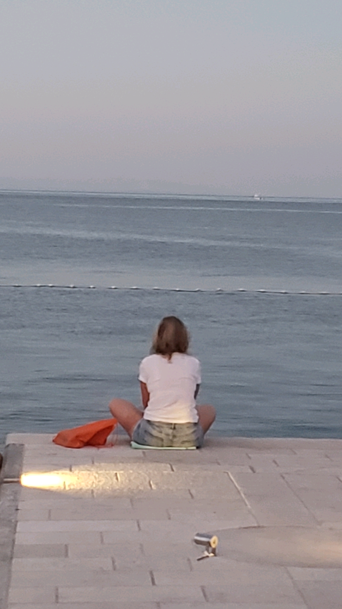 A woman with blonde hair, facing away from the camera, sits cross-legged on a paved area overlooking a body of water. She is wearing a white t-shirt and denim shorts. An orange bag lies to her left. In the distance, the water is calm, with a faint horizon line and a small white object, possibly a boat, visible on the water. A light fixture illuminates the paved surface in the foreground, casting a bright glow and a shadow. Another small object, resembling a flashlight, rests on the pavement. The scene suggests an outdoor setting, likely a promenade or pier in Općina Tučepi, Croatia, given the distinctive paved surface and the proximity to the sea.