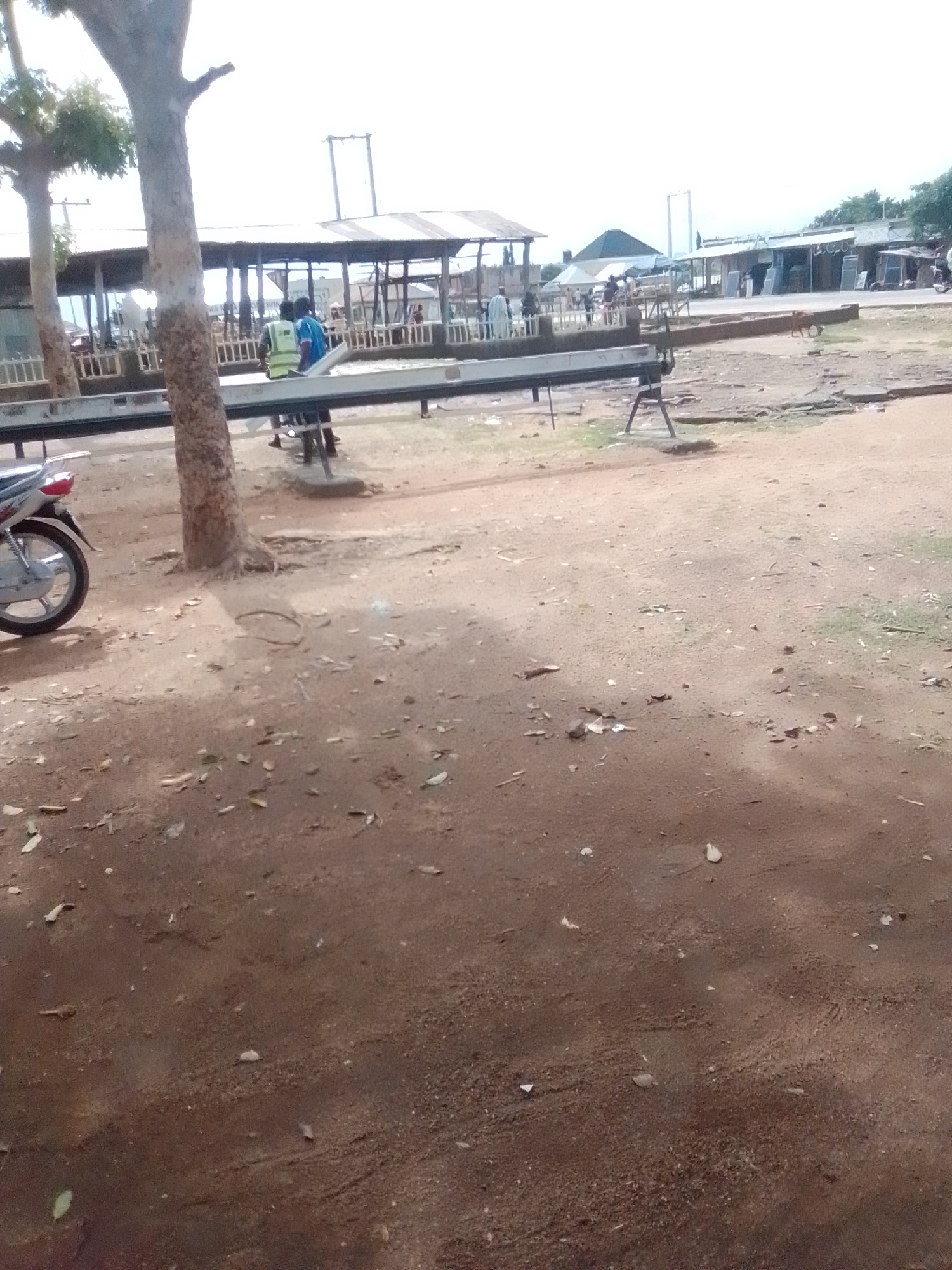 The image depicts an outdoor market scene in Gombe, Nigeria. In the foreground, a dirt ground is scattered with leaves and small debris. A portion of a motorcycle is visible on the left. In the mid-ground, a covered market stall structure runs horizontally across the image. Several people are present within and around the stalls. Two individuals are standing near the left side of the market structure, one wearing a reflective vest. The market stalls appear to be lined with wooden barriers. Further in the background, additional market structures and a tall, slender tower-like object are visible. The sky is overcast and bright.