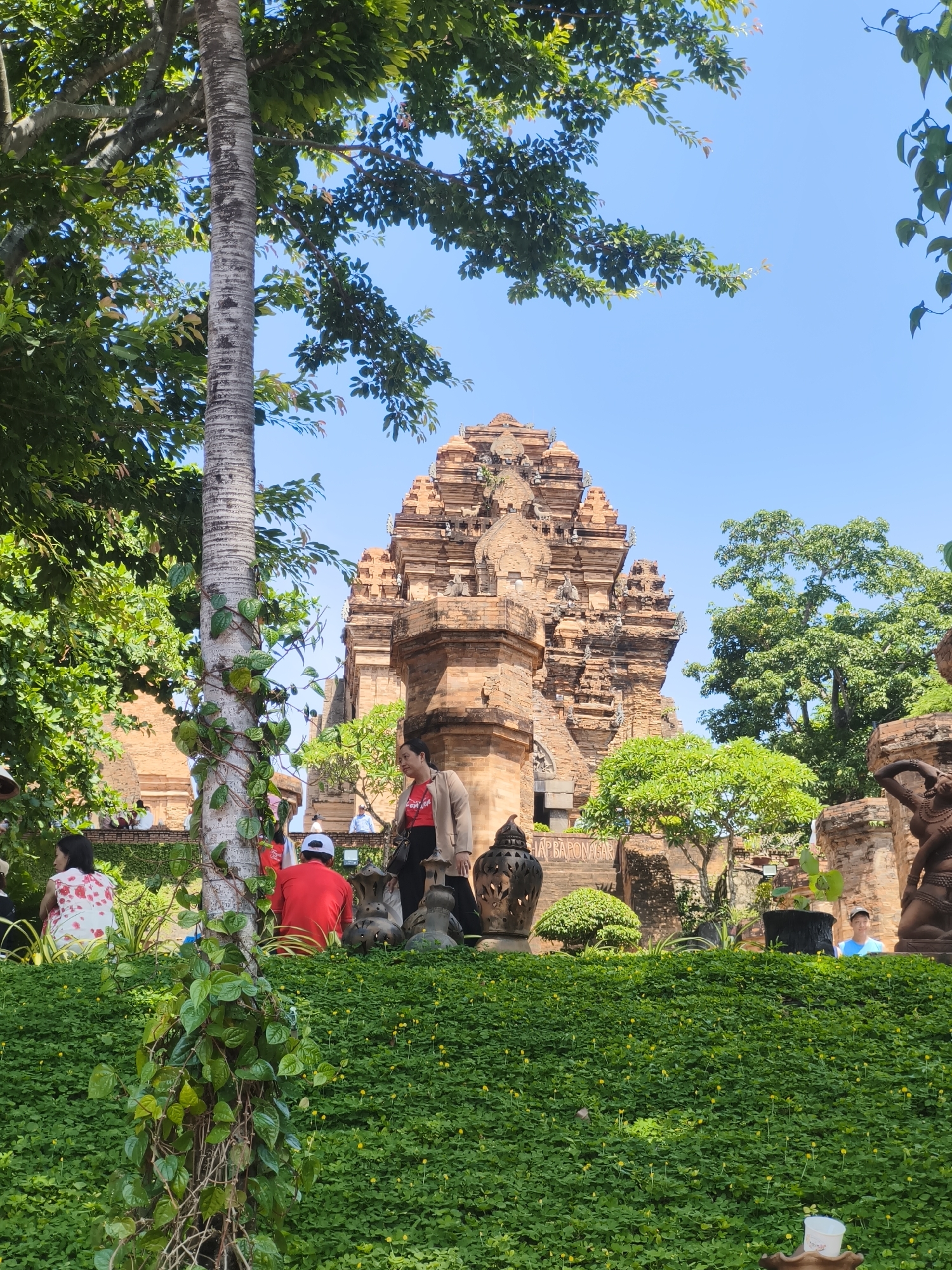 The image depicts the Po Nagar Cham Towers in Nha Trang, Vietnam. In the foreground, a lush green landscape with small yellow flowers and a thick vine-covered tree trunk is visible. Several people are scattered throughout the scene. On the left, a woman in a white and red floral dress is seated. To her right, a person in a red shirt and white cap is also seated. A woman in a beige jacket and red shirt stands near them. In the background, the prominent brick-red Cham towers rise against a clear blue sky. Other people are visible in the distance, some seated along a low wall and others standing. The architecture of the towers is detailed and weathered, suggesting historical significance. Trees with dense green foliage frame the towers and the overall scene.