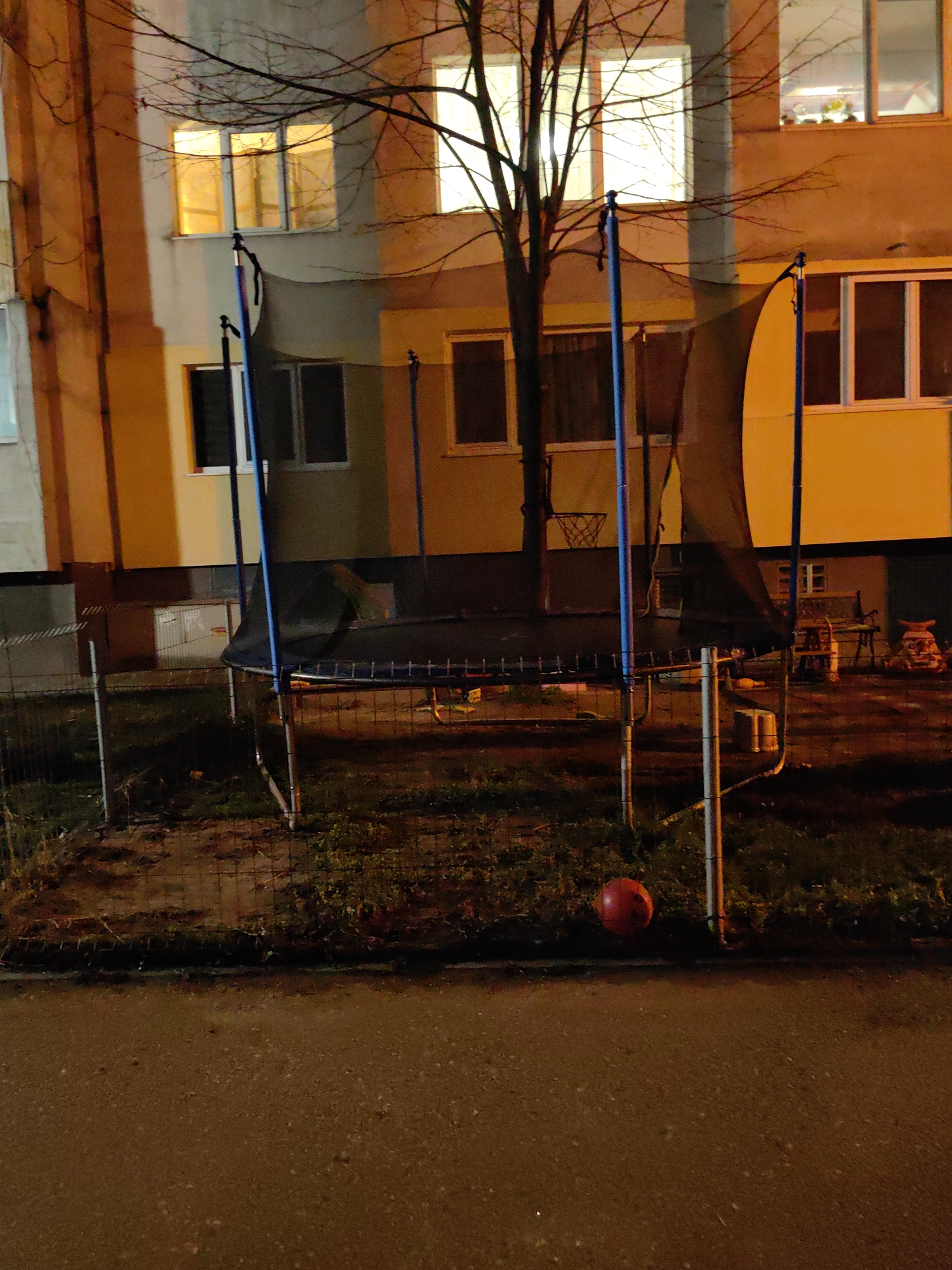 The image shows a trampoline with a safety net in a courtyard at night. The trampoline is surrounded by a fence. A red ball is on the ground near the trampoline. In the background, there is a building with lit windows and bare trees. The location is likely in Sofia, Bulgaria, as indicated by the context of the prompt. There are no people visible in the image.