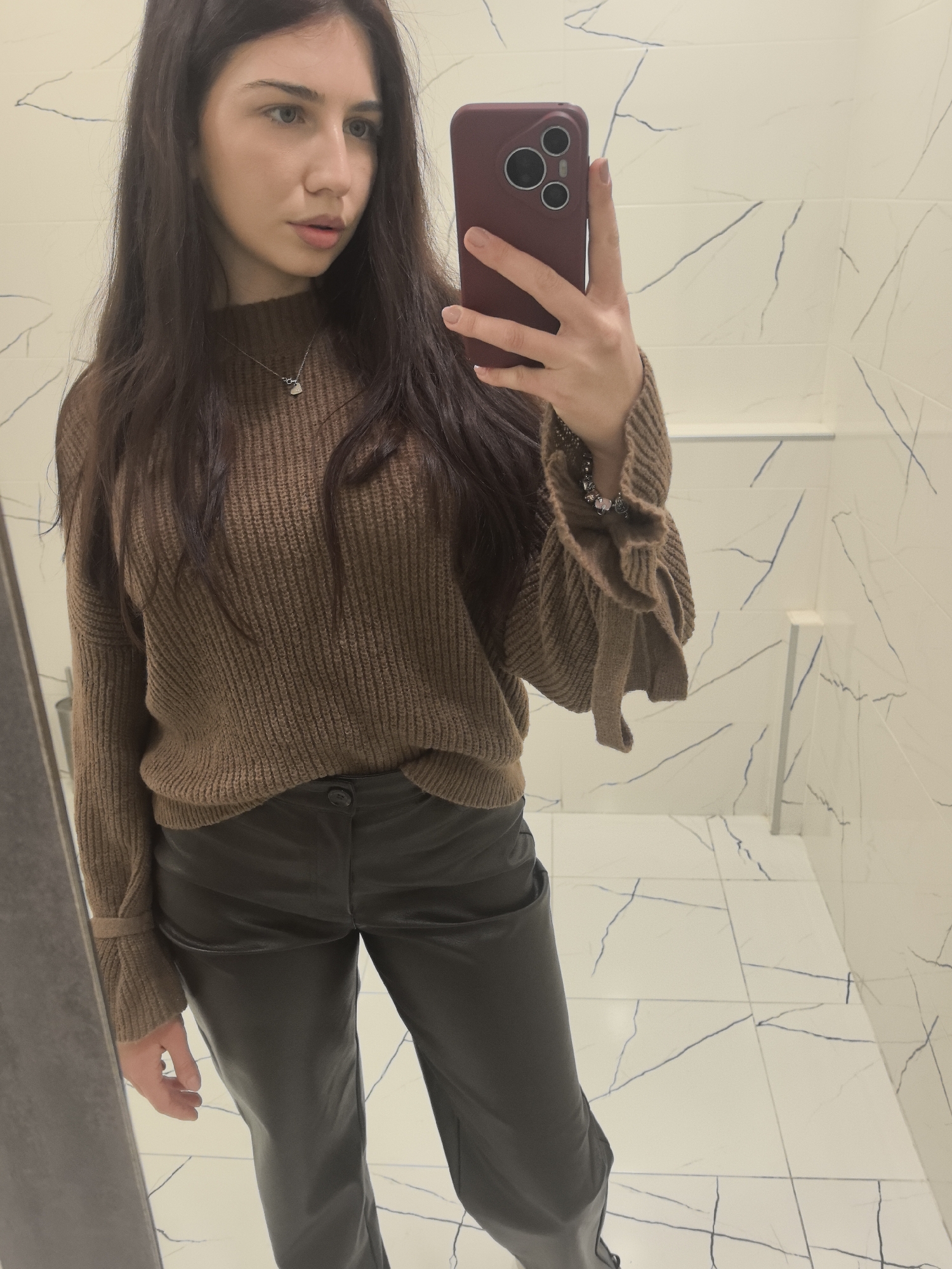 A woman is taking a mirror selfie indoors. She is wearing a brown sweater with ruffled sleeves, black pants, and a necklace. She is holding a phone with a burgundy case. The background is a tiled wall with marble-like patterns and white tiled flooring. The location is potentially in a building in Stara Zagora, Bulgaria, but there is no direct visual evidence in the image to confirm this.