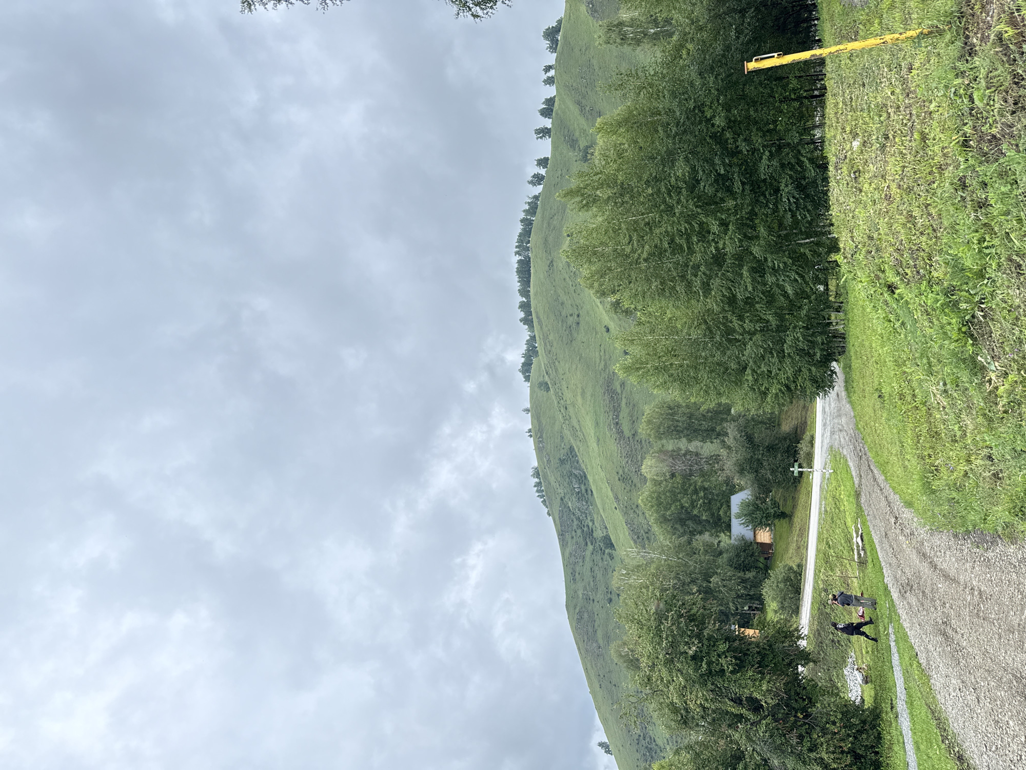 The image depicts a rural landscape with a prominent green hill under a cloudy sky. In the foreground, a gravel path curves through grassy terrain. Two individuals are visible on the path near a cluster of trees. To the left, a small wooden building is nestled among the trees, and further back, a paved road is visible. A white cross stands beside the road. The scene is set in Нижнекаянча, Russia.