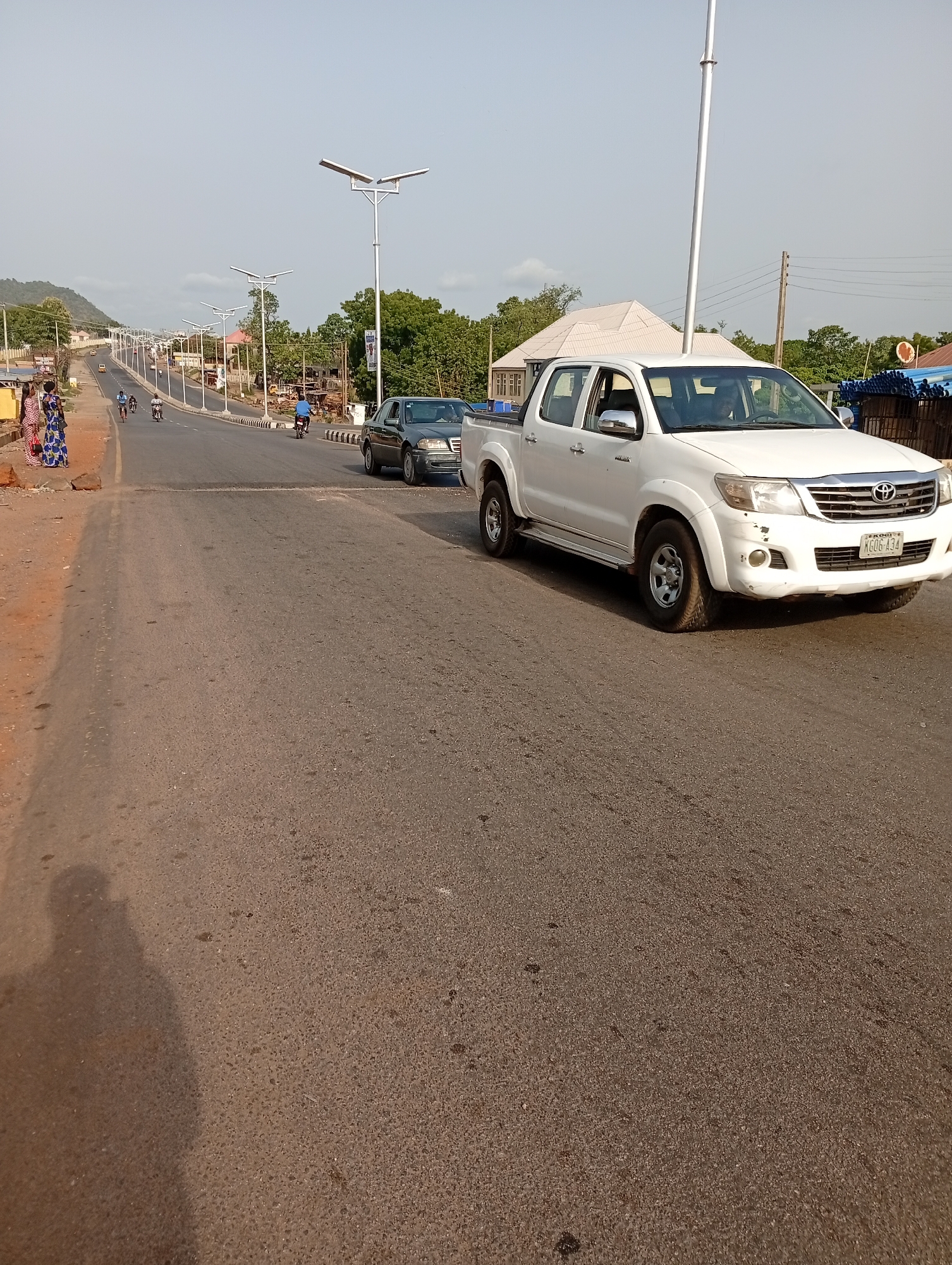 The scene depicts a road in Zango-ndaji, Nigeria. A white Toyota Hilux pickup truck is parked on the right side of the road. A dark-colored sedan is further down the road, and several people are walking along the left side of the road in the distance. Three people on motorcycles are seen. Streetlights line both sides of the road. The road appears to be paved. A hill is visible in the background.