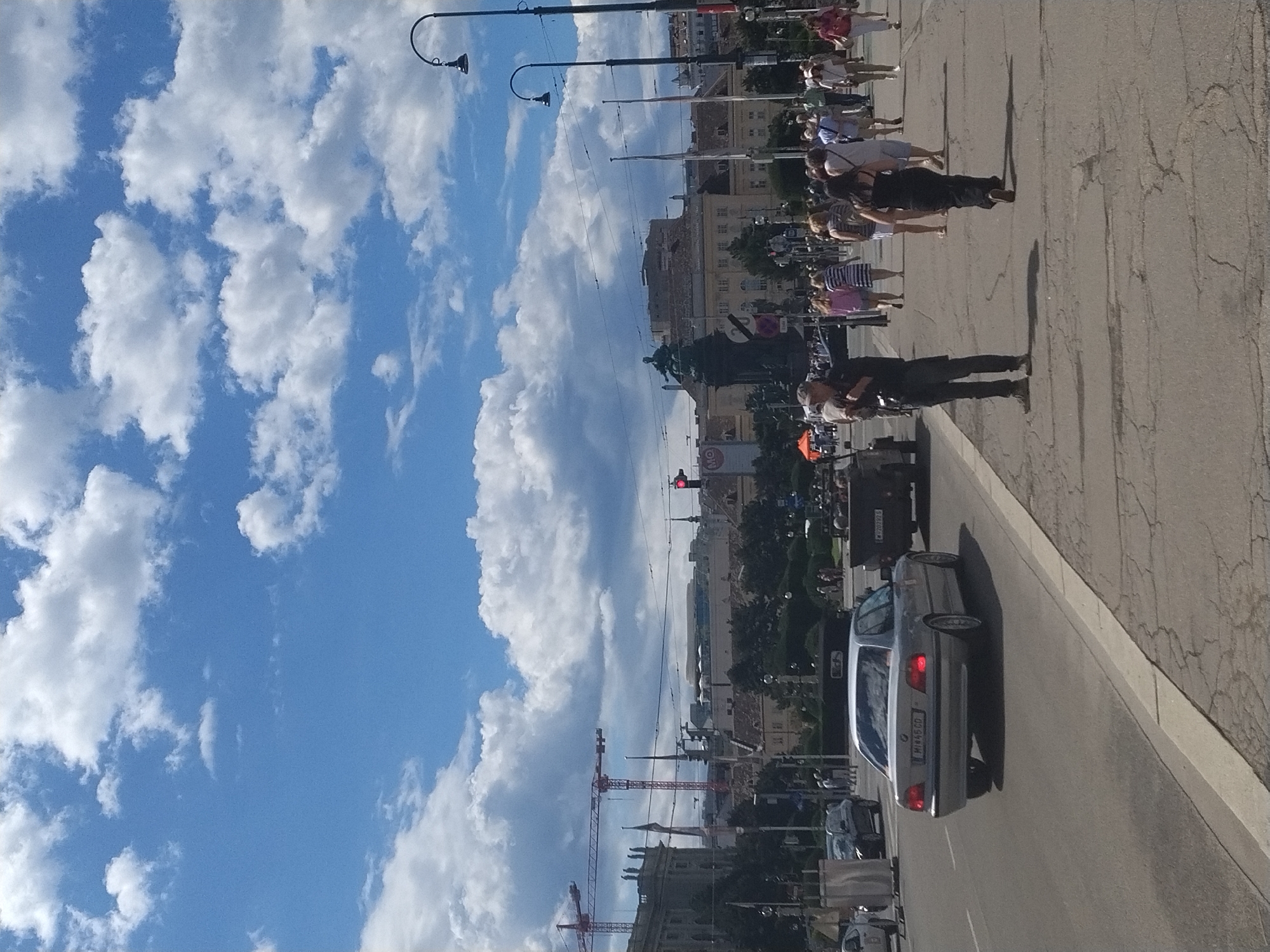 The image depicts a sunny day in Vienna, Austria, likely near the Heldenplatz. A man in a dark shirt and pants stands facing right, looking towards the right side of the frame with his right arm extended. He is on a paved sidewalk adjacent to a street. Several cars are visible on the street, including a silver sedan in the foreground and a dark military-style vehicle behind it. A crowd of people, dressed in summer attire, are walking on the sidewalk. In the background, buildings with classical architecture are visible, along with several tall construction cranes and flags. The sky is a vibrant blue with scattered white clouds. The scene captures a lively urban environment with elements of both historical significance and modern activity.
