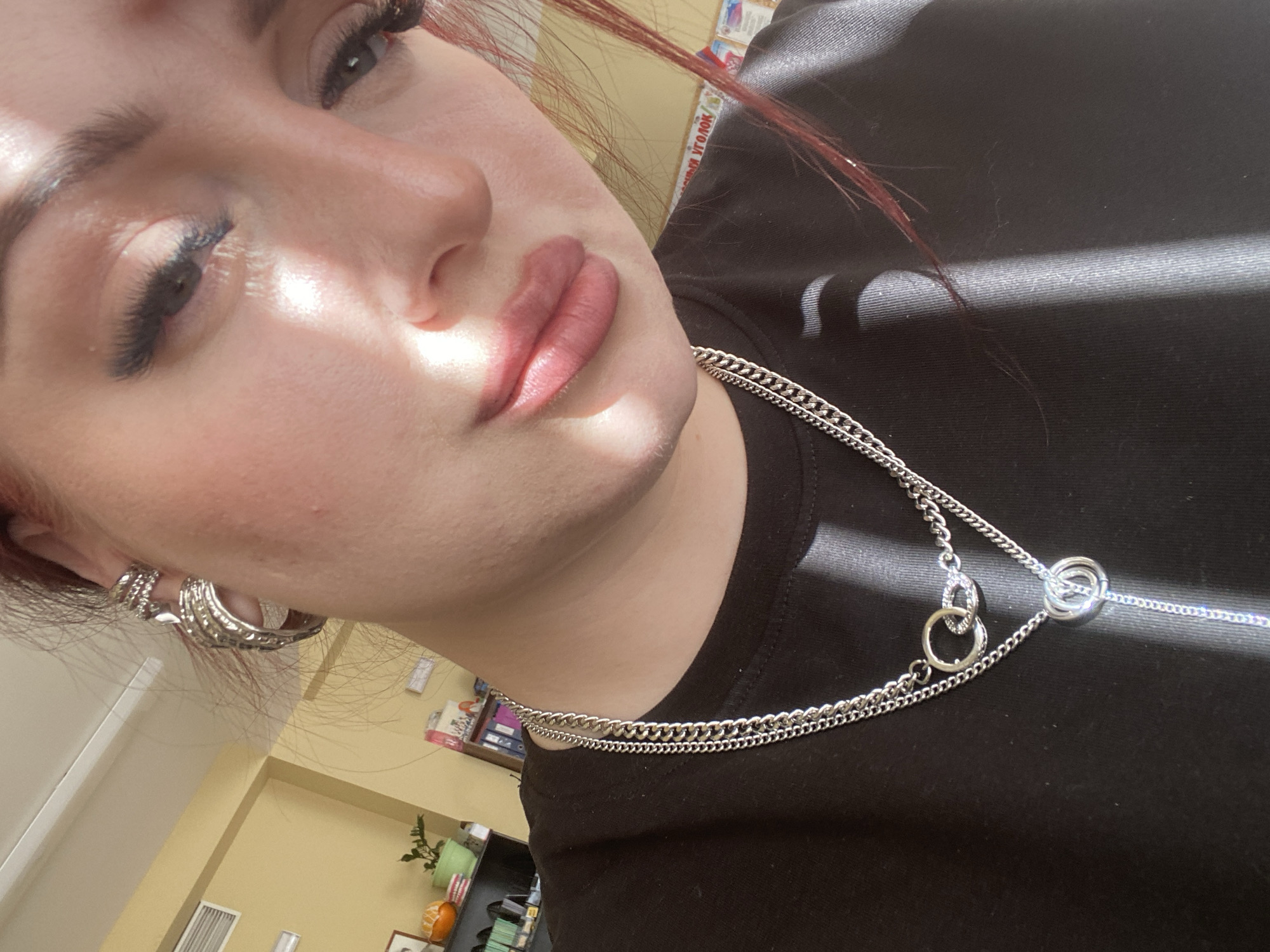 A close-up, low-angle shot of a woman with fair skin, red-tinted hair, and defined eyebrows, positioned against a background suggesting an indoor setting. The woman's face is partially illuminated by sunlight casting shadows across her features, particularly on her cheek and neck. She is wearing a black t-shirt and a silver layered necklace with ring-shaped accents. Multiple silver hoop earrings adorn her left ear. The background includes a glimpse of a yellow wall and office supplies, potentially indicating a room in the city of Chaikovsky, Russia. The focus is on the woman's face and jewelry, with the lighting creating stark contrasts.