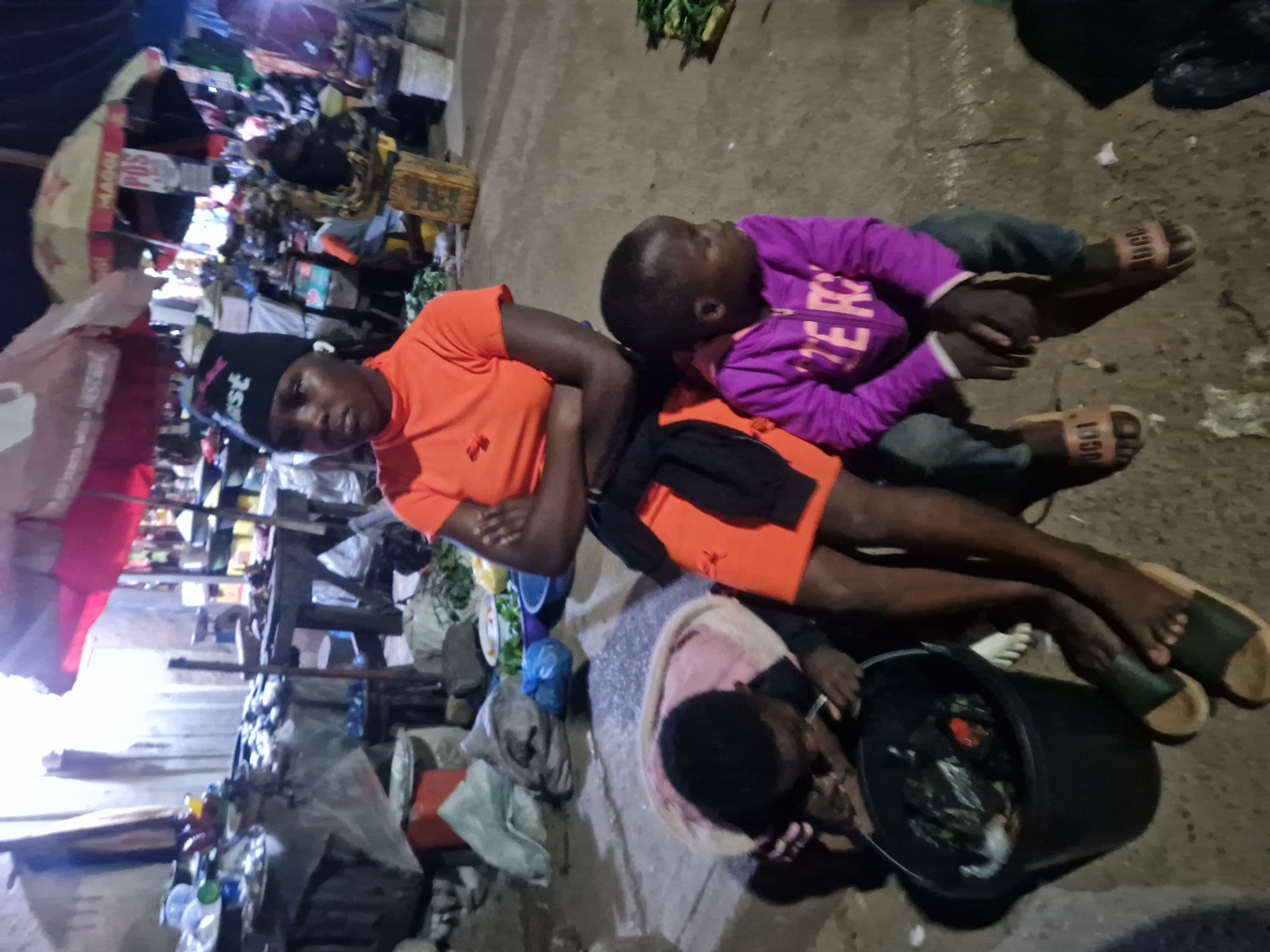 In Kabong, Nigeria, an outdoor market scene unfolds at night. A young woman, wearing an orange long-sleeved shirt and a black beanie, sits with her arms crossed, her gaze directed forward. Beside her, a young child in a purple hooded jacket and jeans looks to the right. To the left, another child, wearing a pink and grey hooded garment, leans over a black bucket filled with items, seemingly examining its contents. The ground beneath them is a mix of concrete and dirt, with debris scattered around. In the background, market stalls are visible, some with umbrellas providing shade, and various goods are displayed. The lighting suggests artificial illumination from the market or surrounding structures.