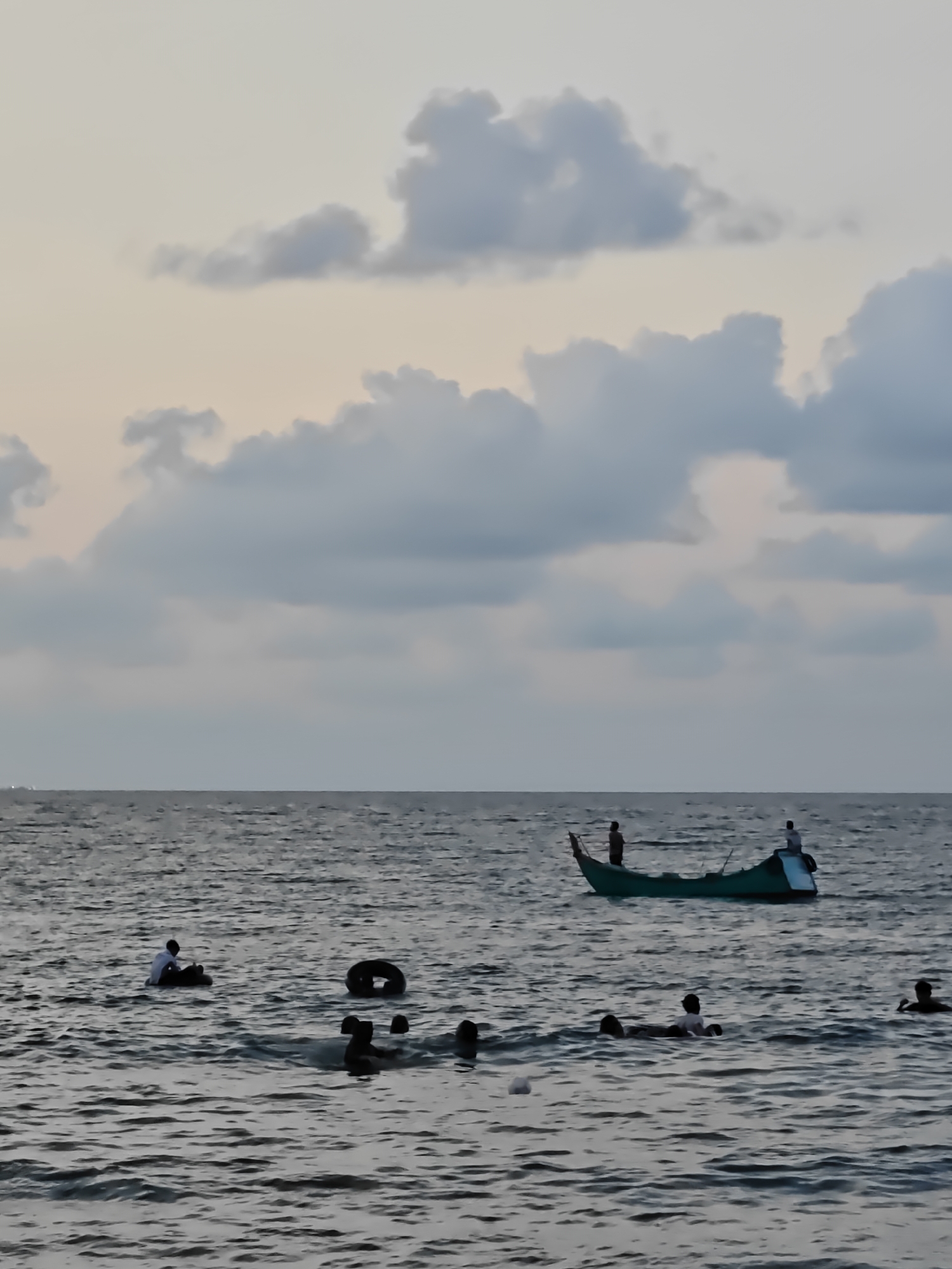 The image depicts a seascape in Lhokseumawe, Indonesia, at dusk. The sky is filled with gray and white clouds. The ocean surface is choppy, with visible waves. In the foreground, several people are swimming in the water. Some are floating on inner tubes, while others are swimming freely. Further out, a small green fishing boat is visible, with two figures standing on board. The boat is oriented toward the right side of the frame. The scene suggests recreational activity near the shore.