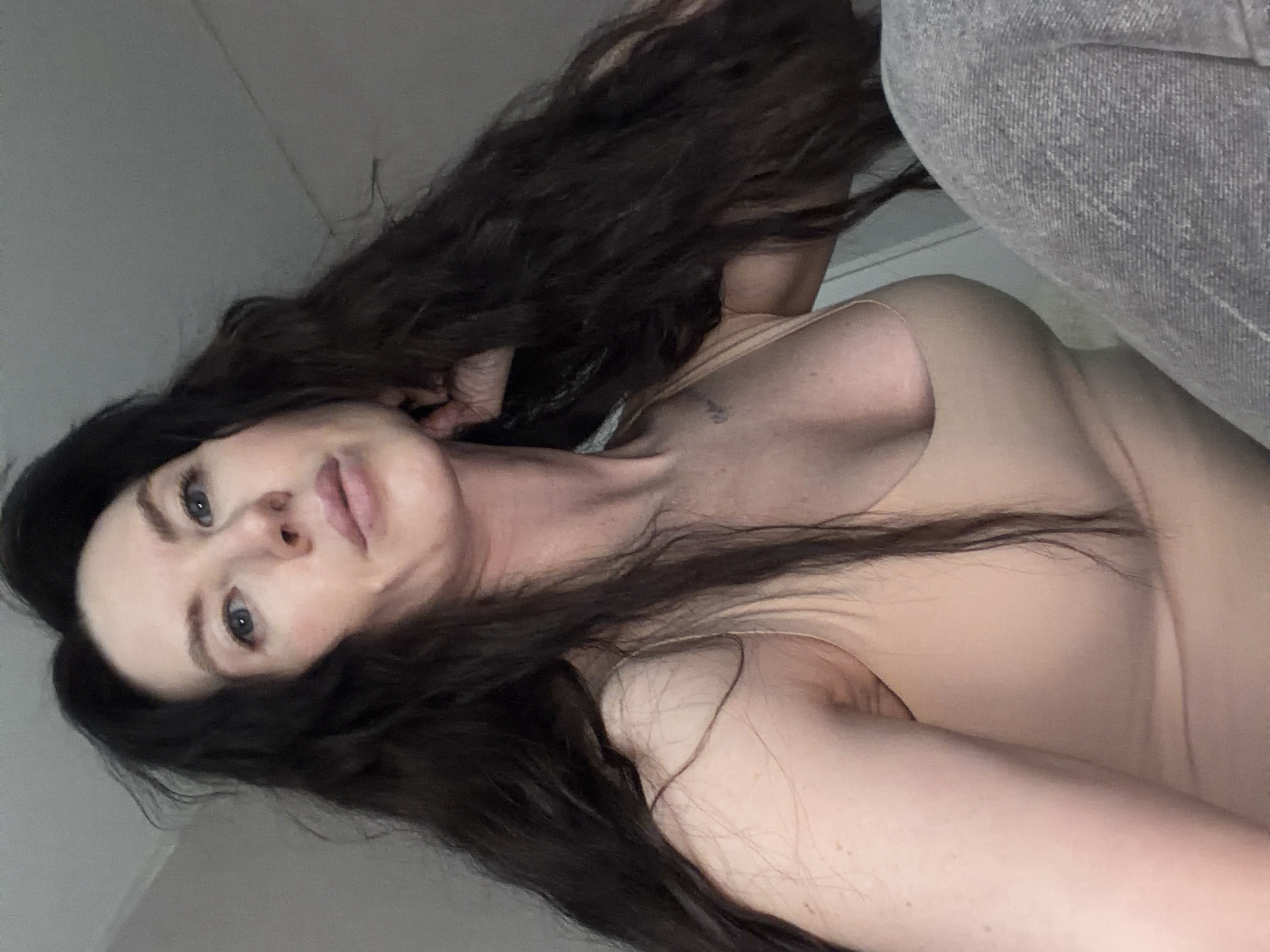 An adult female is depicted in a low-angle indoor portrait. She possesses long, dark, wavy hair, blue eyes, and full lips. Her complexion is fair. She is clad in a light beige or nude-colored form-fitting tank top or bodysuit. A small, dark tattoo is visible on her upper left chest/collarbone area. Her right hand is partially obscured, appearing to rest near the back of her head or neck. The lower right portion of the image shows grey fabric, consistent with trousers. The background consists of a plain, light-colored wall and ceiling, offering no specific identifying features. The precise location within Perm, Russia, cannot be determined from the provided visual information.