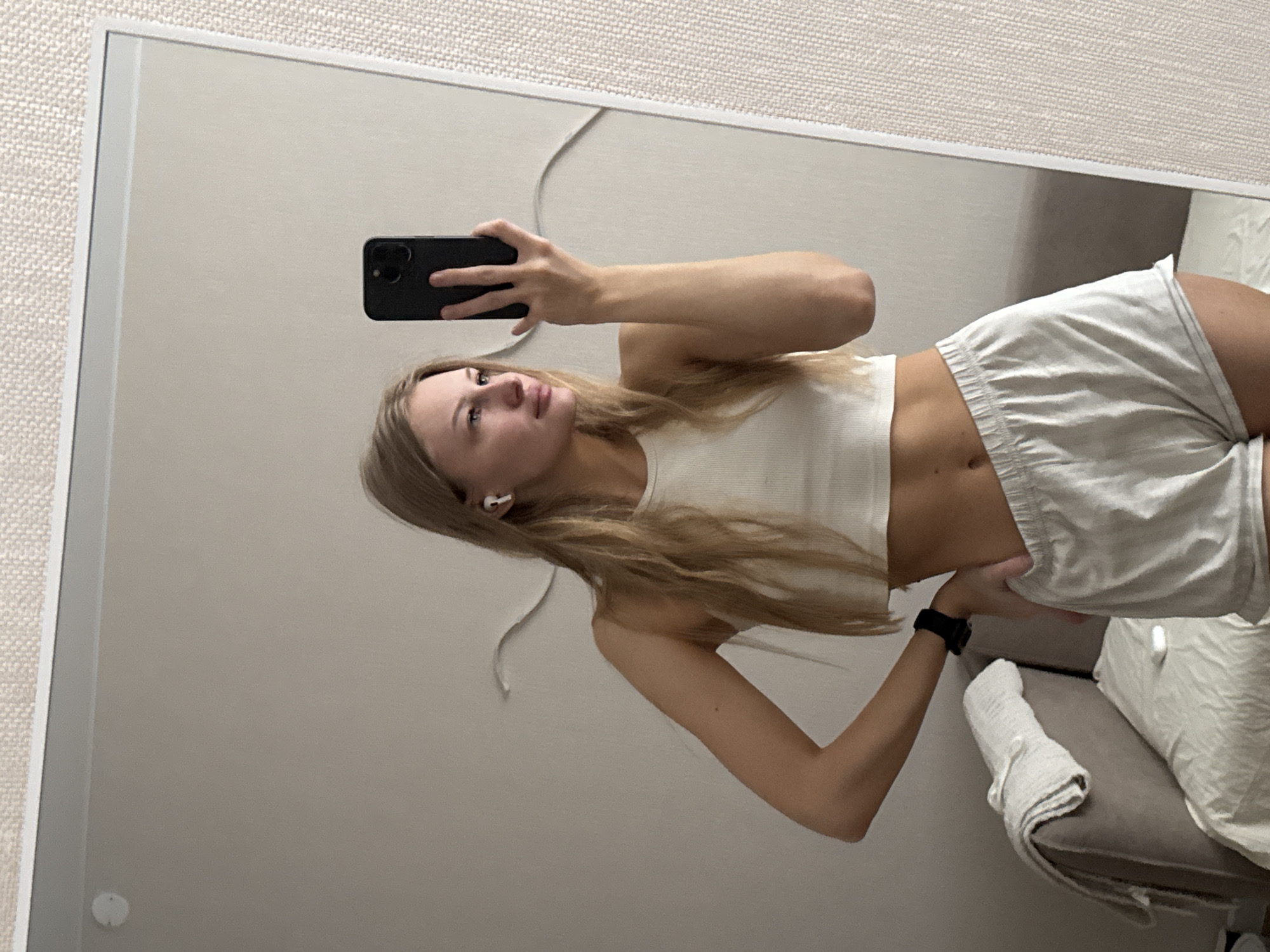 A young woman with blonde hair is taking a mirror selfie in a room. She is wearing a white tank top and light-colored shorts. She has on white wireless earbuds and a black watch. She is standing with her left hand on her hip and her right hand holding a black smartphone. In the background, there is a sofa with a blanket draped over it. The wall behind her appears to be textured. There is no indication of the location being in the city of Perm, Russia.