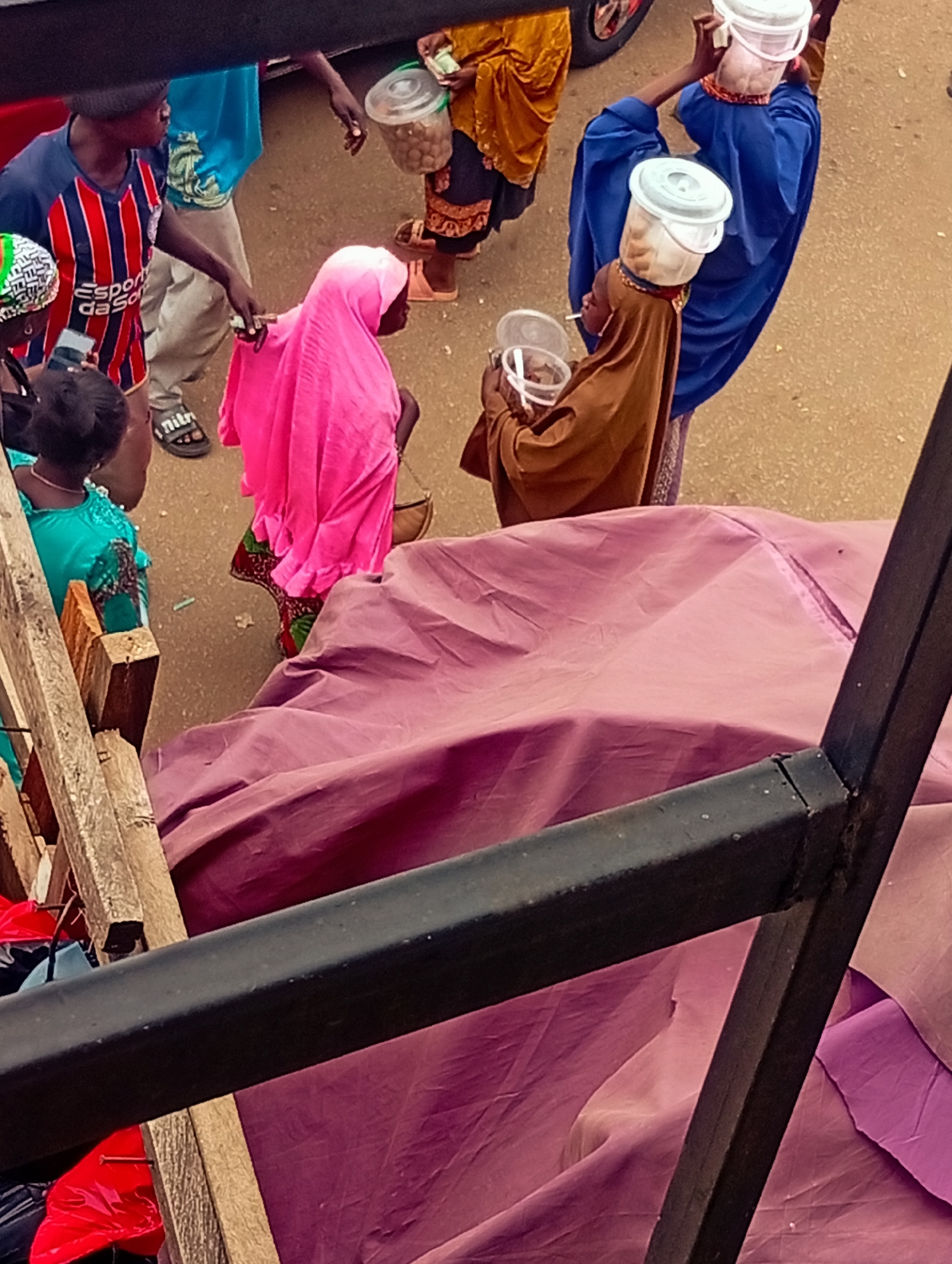 Captured from an elevated perspective, the photograph presents a street scene. People are visible, some carrying containers on their heads. The attire of the individuals varies, including traditional robes and modern clothing. Transactions appear to be taking place. The environment is urban. The location of the city is Unknown, Unknown.