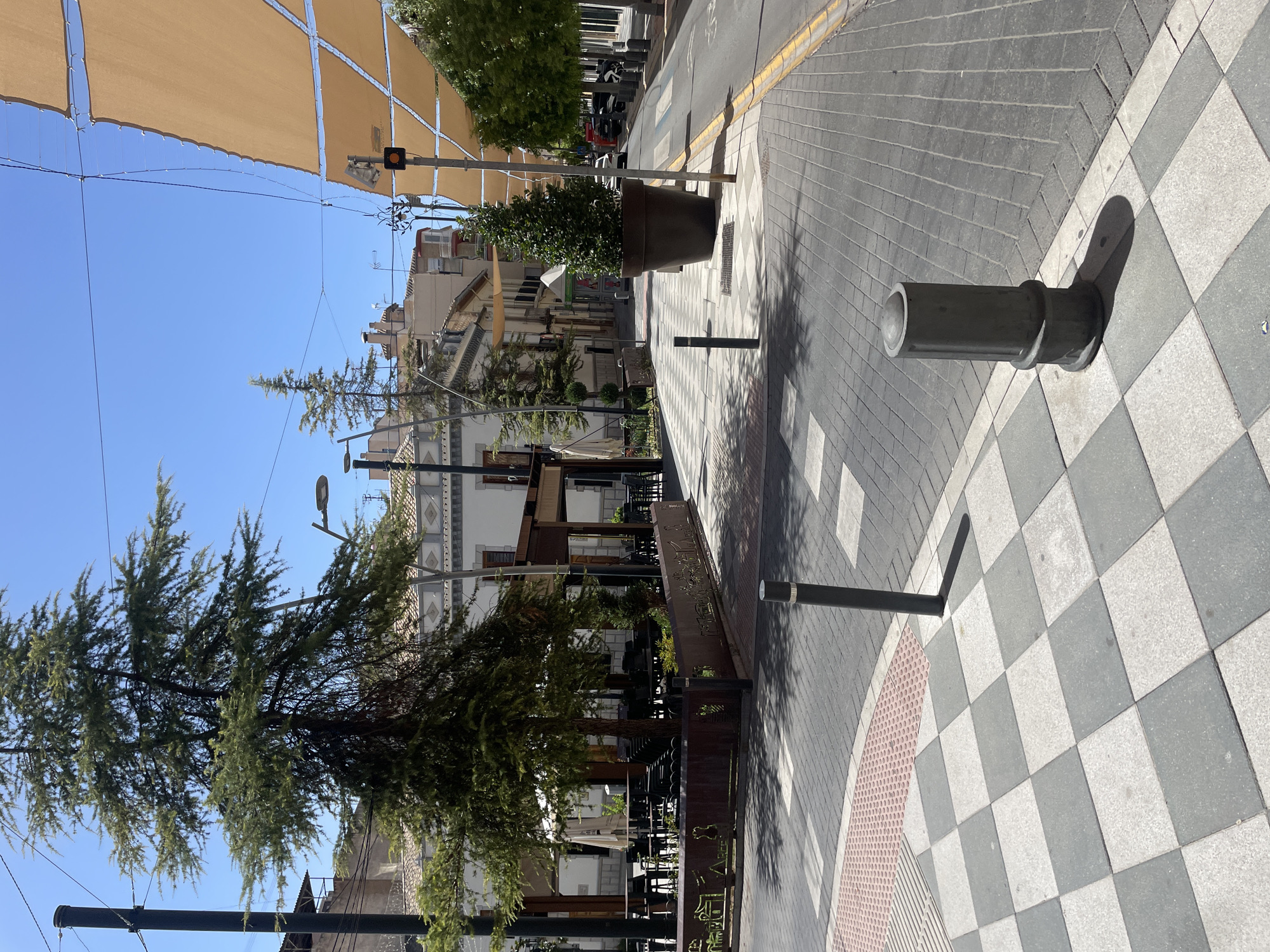 The image depicts a sunlit pedestrian area in Atarfe, Spain. In the foreground, a checkered pattern of light grey and dark grey square tiles forms a pathway. A dark grey, cylindrical bollard stands on the tiles, casting a shadow. Further into the scene, the checkered pattern transitions to a more intricate pattern of light grey and white tiles with diagonal lines and small square inlays, bordered by a reddish-pink textured tactile paving area.
A dark grey cylindrical bollard is positioned at the corner where the checkered tiles meet the patterned tiles. Beyond this, a paved road with grey brickwork and a yellow painted border runs along the right side of the image. Several parked vehicles, including a white car and motorcycles, are visible on the right side of the road. A tall, dark brown planter containing a dark green conical tree stands on the patterned tiles, situated near the edge of the road.
To the left, a shaded outdoor seating area for a cafe or restaurant is visible, with tables and chairs. A large tree with dark green foliage stands behind a decorative metal barrier in front of the seating area. Buildings with white facades and tiled roofs line the street in the background. Light brown fabric awnings are stretched across the street, providing shade. The sky is clear and blue. Power lines are visible strung across the upper portion of the image.