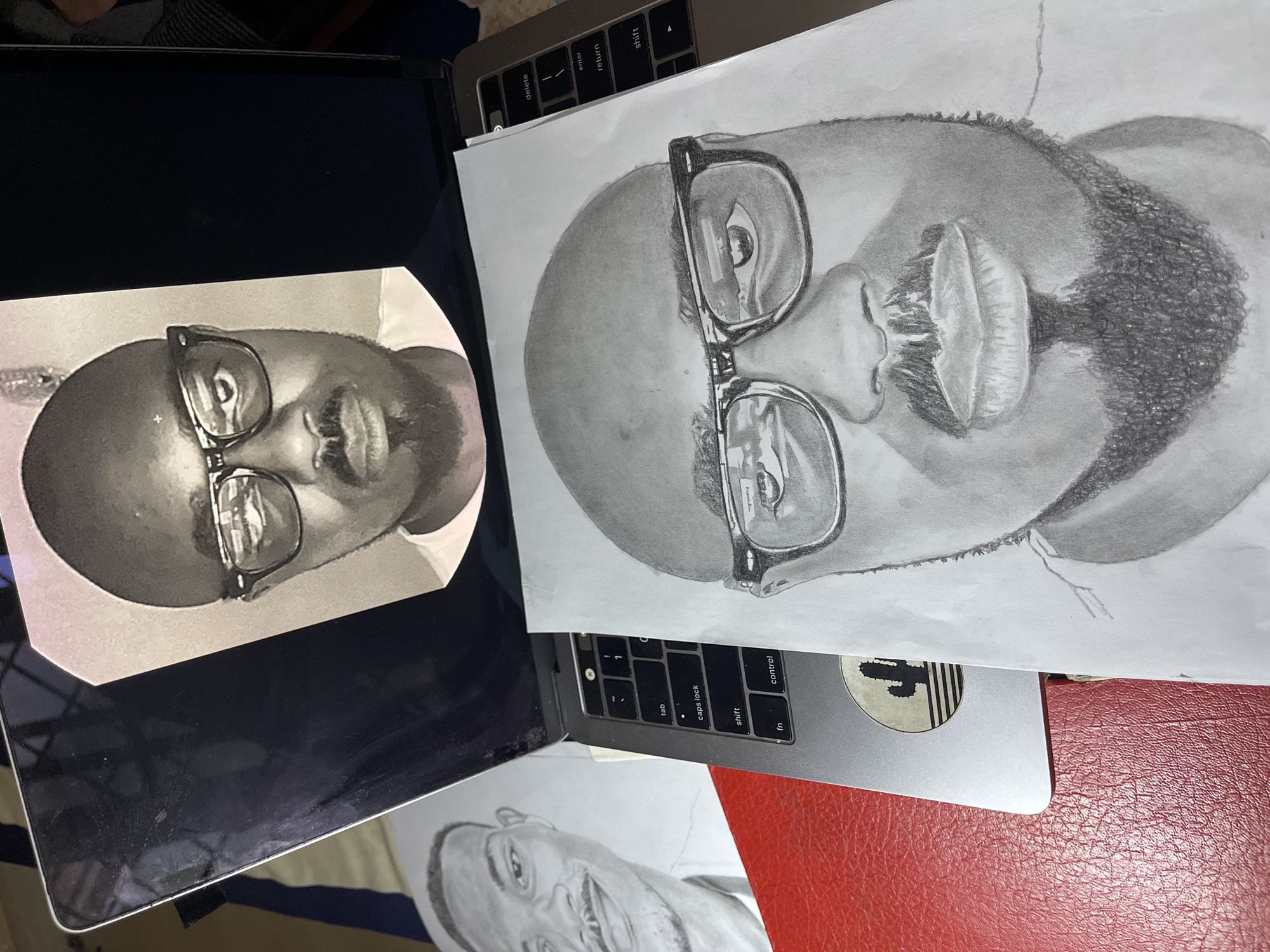 The image shows two pencil drawings of a bald man with a beard and glasses. One drawing is on a piece of paper resting on a laptop keyboard. The other drawing is on a separate piece of paper, positioned on the laptop screen. There is a third drawing of the same man visible at the bottom of the image. A red leather surface is visible to the right of the laptop. A sticker of a cactus is on the laptop. The location is unspecified. The city and country are not apparent from the image.
