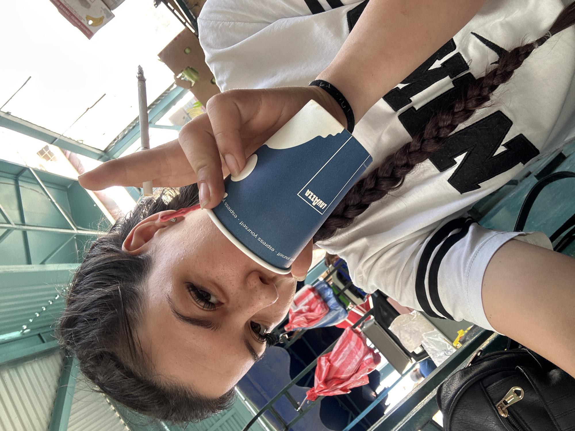 The image shows a young woman with dark hair, holding a coffee cup and a cigarette. She is wearing a white t-shirt with black stripes on the sleeves. The background suggests an outdoor market or a covered seating area, with green metal structures and bags hanging in the background. The woman is looking at the camera, and her left hand is raised, holding the cigarette between her index and middle fingers. The coffee cup has the Lavazza logo on it. The overall setting appears to be casual and informal. No specific location in Sliven, Bulgaria is identifiable from the image.