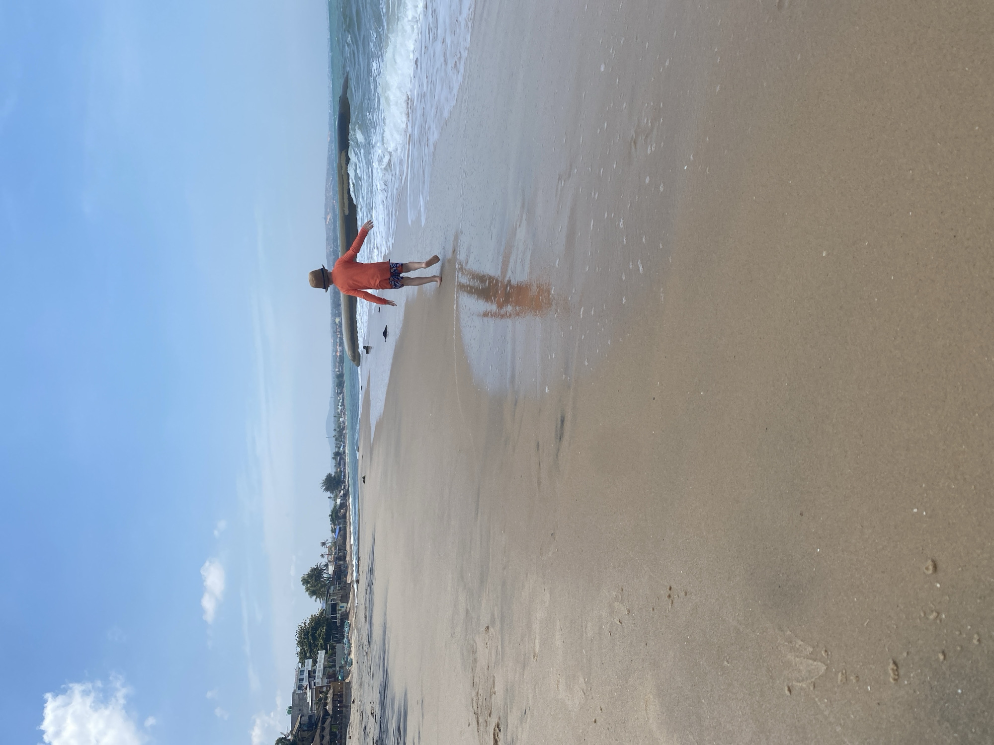 The image depicts a person standing on a sandy beach in Phu Thuy Ward, Vietnam. The person is wearing an orange long-sleeved shirt and patterned shorts, facing away from the viewer towards the ocean. The beach is wet from the receding waves, creating a reflective surface. A dark object lies on the shoreline, near the person's feet. The background shows a glimpse of buildings and vegetation along the coast, under a clear blue sky with some scattered clouds. The beach dominates the lower portion of the image, while the sky occupies the upper portion.