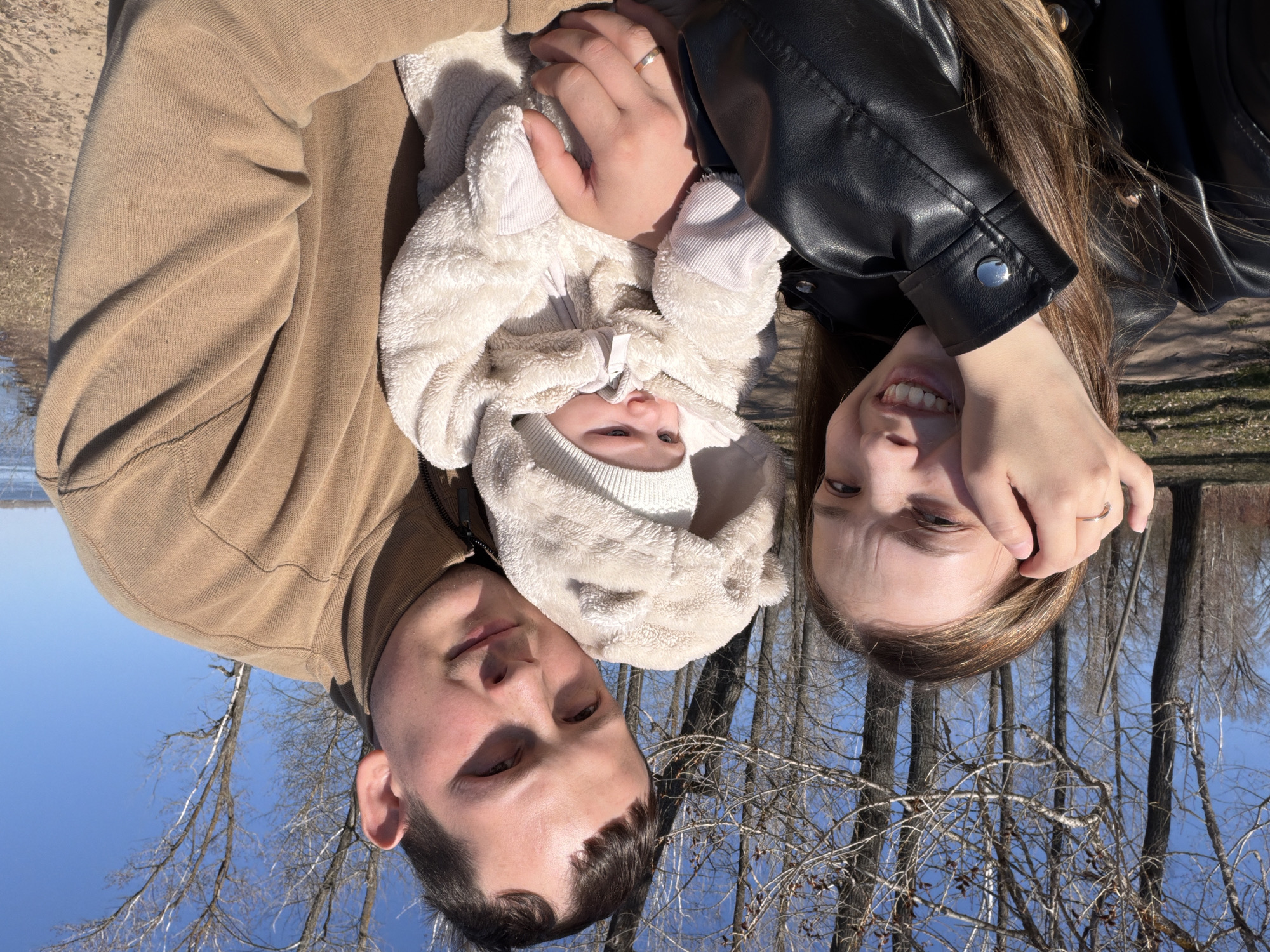 A family of three is photographed in Noviy, Russia, from a point-of-view above them, with the image oriented such that they appear upside down. The family consists of a man, a woman, and a baby. The man is on the left, wearing a tan sweatshirt. The woman is on the right, wearing a black leather jacket. The baby is in the center, bundled in a white hooded snowsuit. The adults are holding the baby. Behind them, bare trees are visible against a blue sky. A body of water and a sandy area are visible in the background.